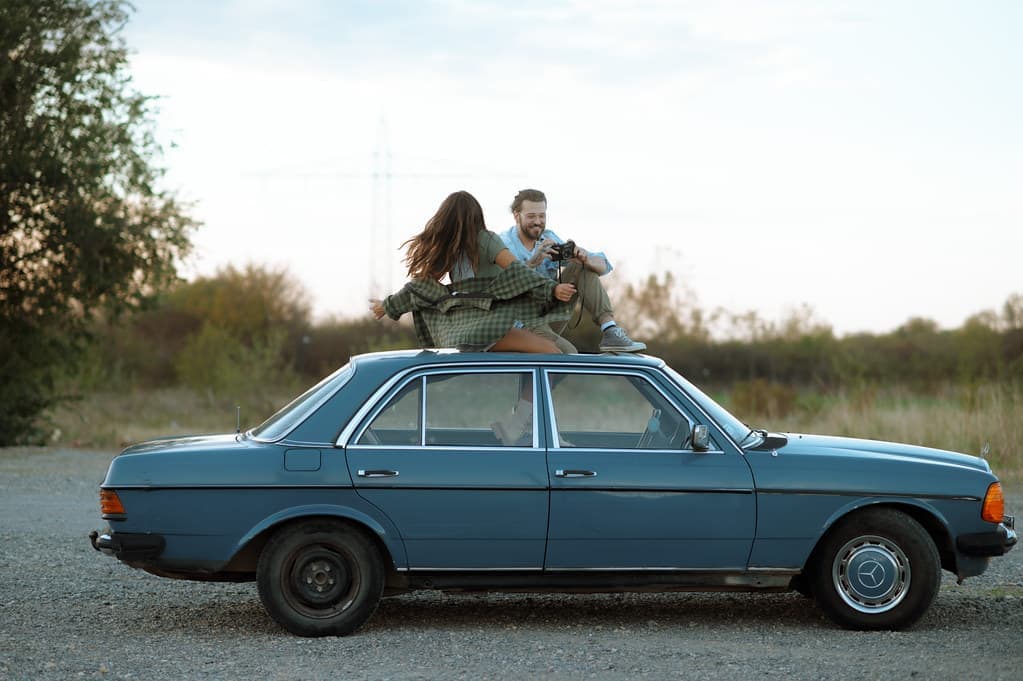 A couple sitting on the roof of their classic blue car at dusk