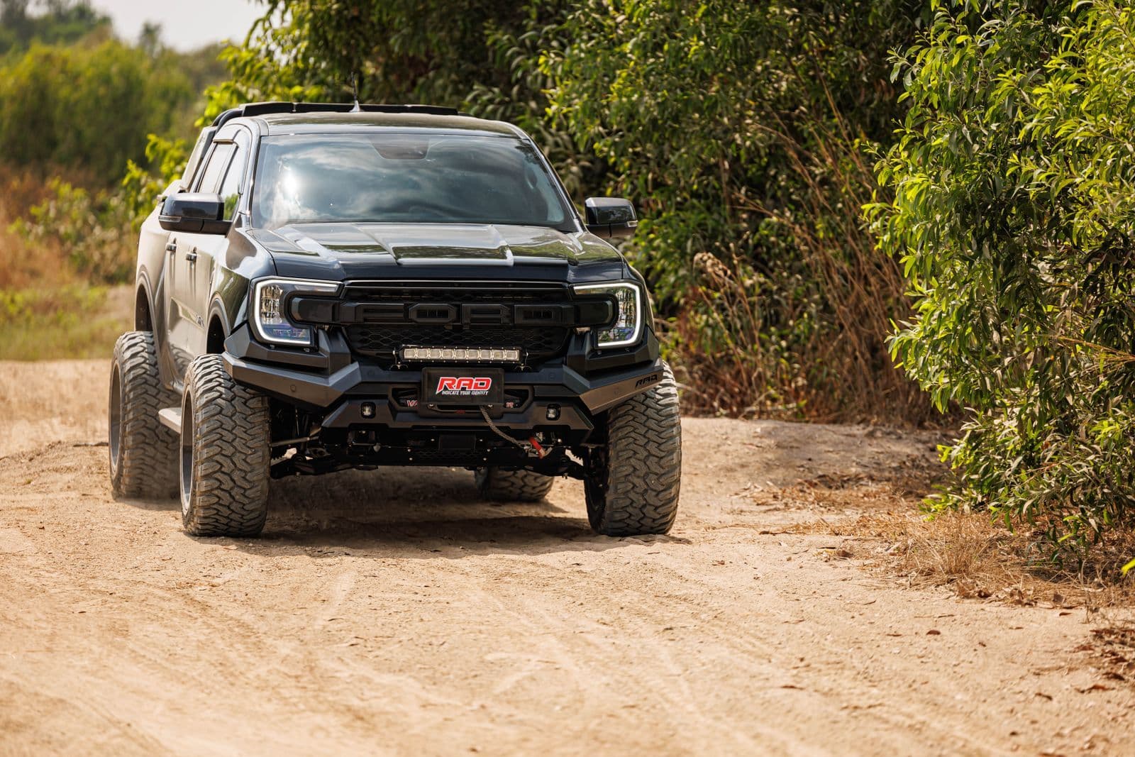 A lifted black dual-cab ute with oversized off-road tyres, a light bar and an aftermarket bumper on a dirt track.