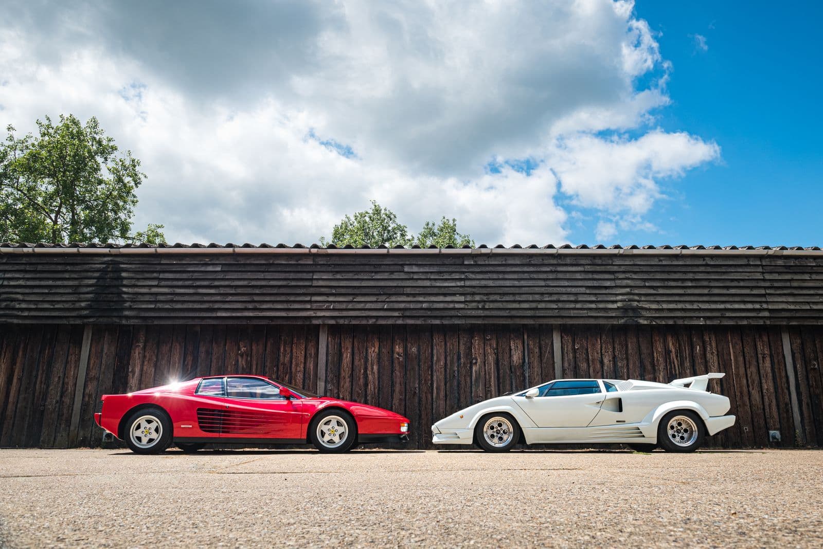 A red Ferrari Testarossa and a white Lamborghini Countach parked nose to nose in front of a timber barn.