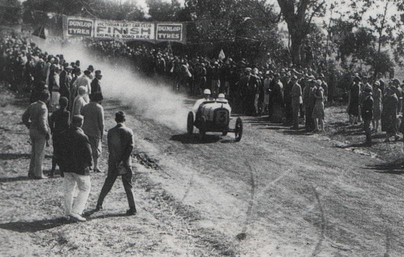 A small open racing car with two occupants on a dirt road, photographed during the 1928 100 Miles Road Race.
