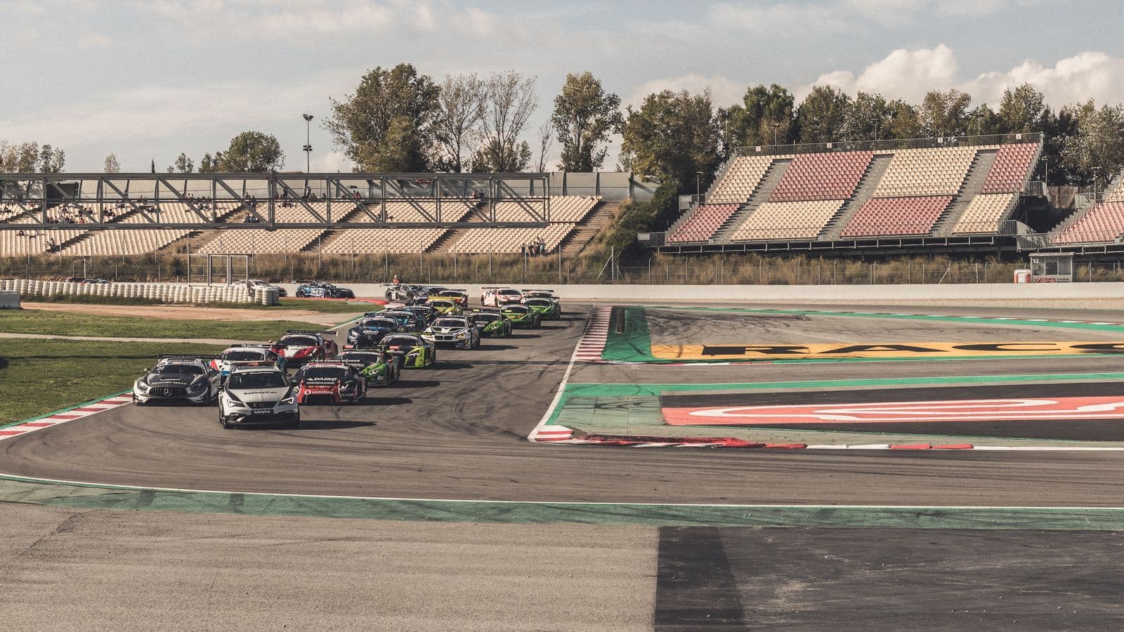 A field of touring cars queued nose to tail on a race circuit beside empty grandstands.