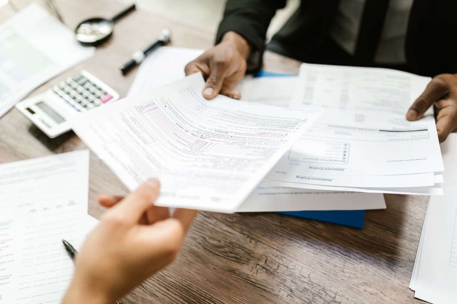 Two people passing printed forms across a wooden desk, with a calculator and pen beside them.