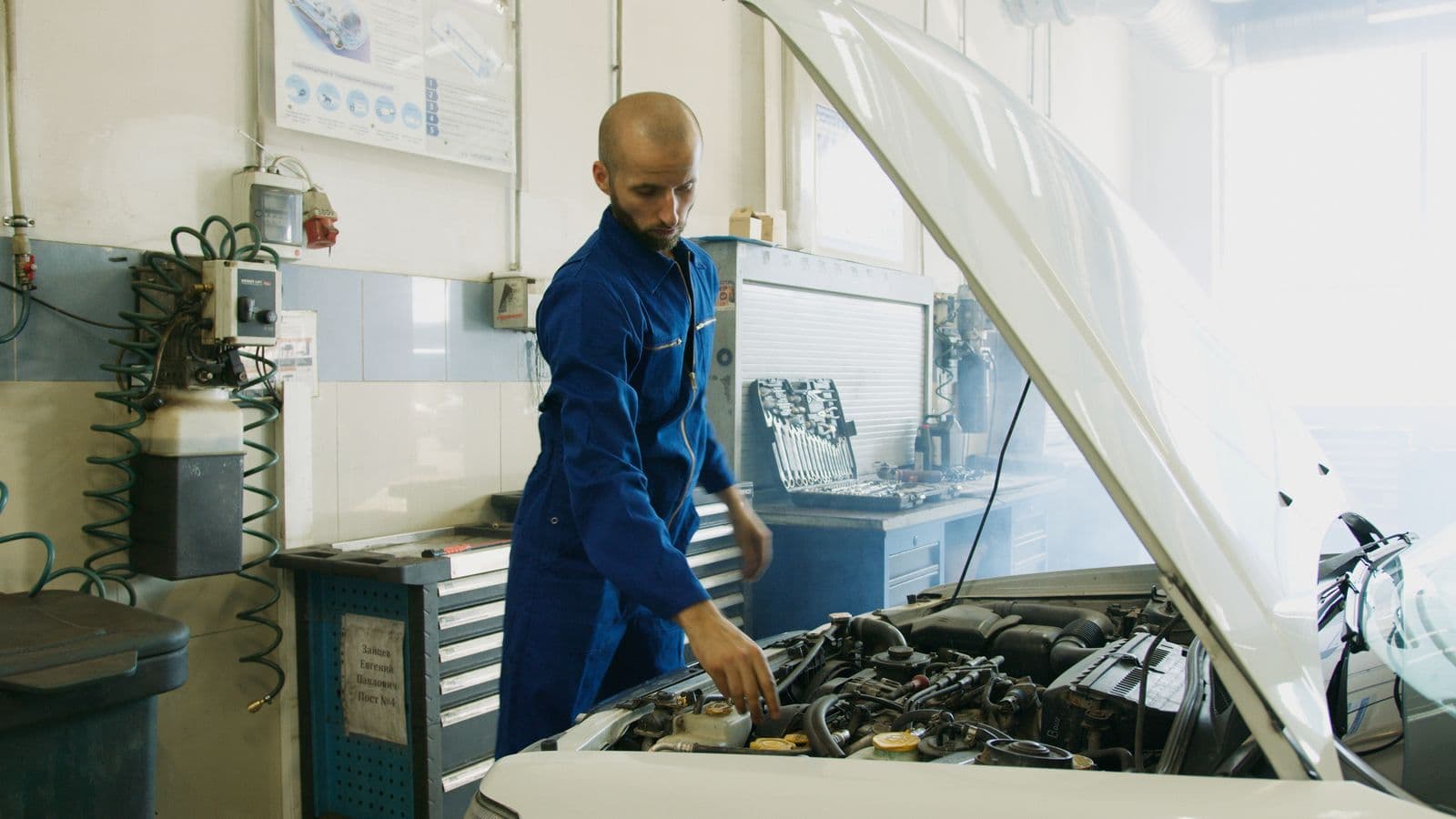 A mechanic in blue overalls leans over the open engine bay of a car in a workshop.