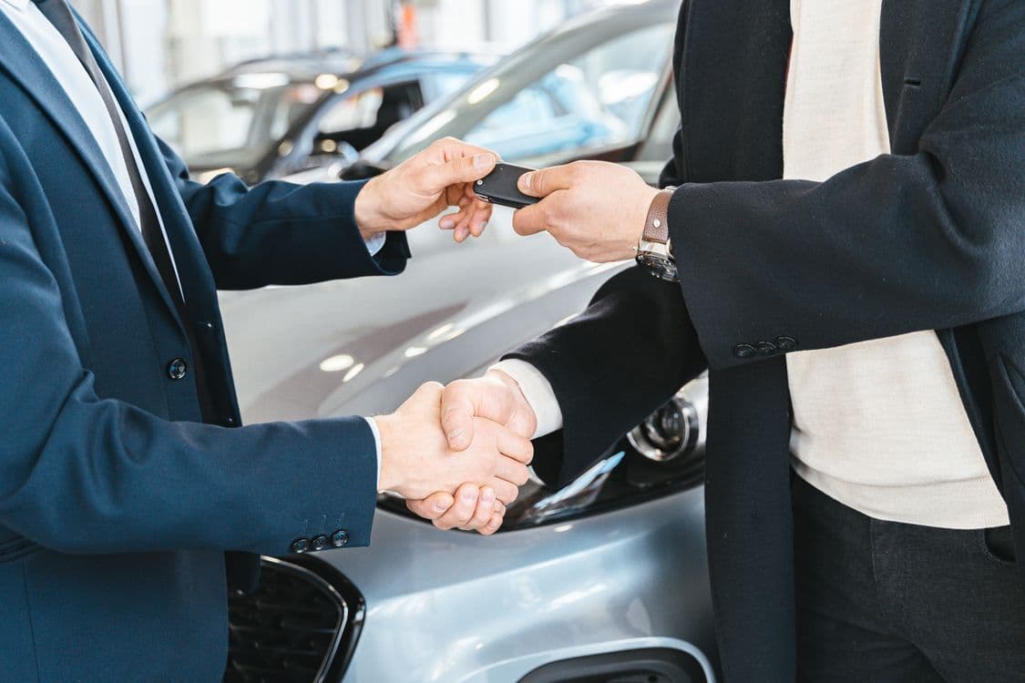 Two people exchange a car key and shake hands in front of a car. Illustrative stock photo.