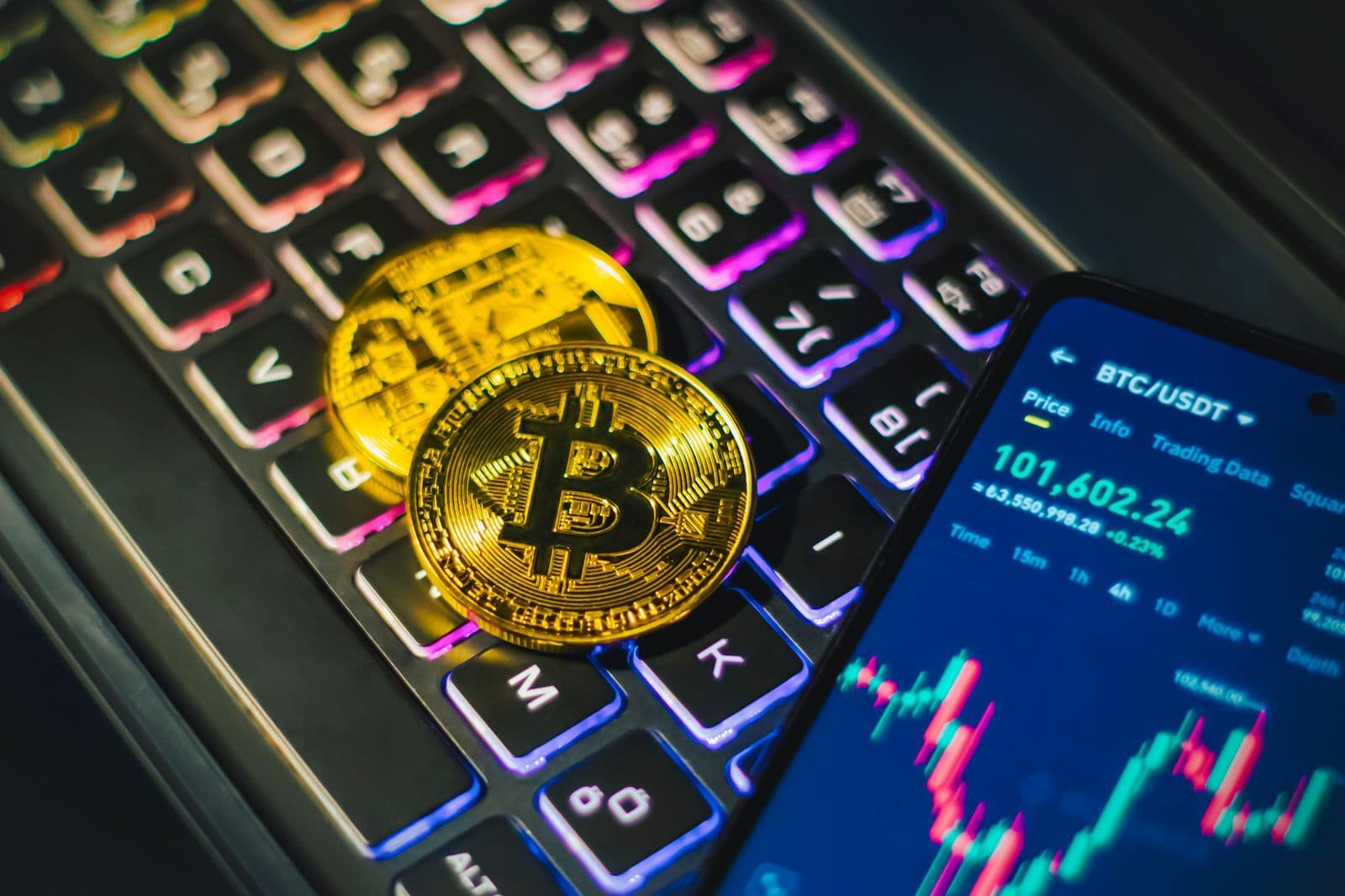 Two gold Bitcoin coins resting on a backlit laptop keyboard beside a phone showing a candlestick price chart.