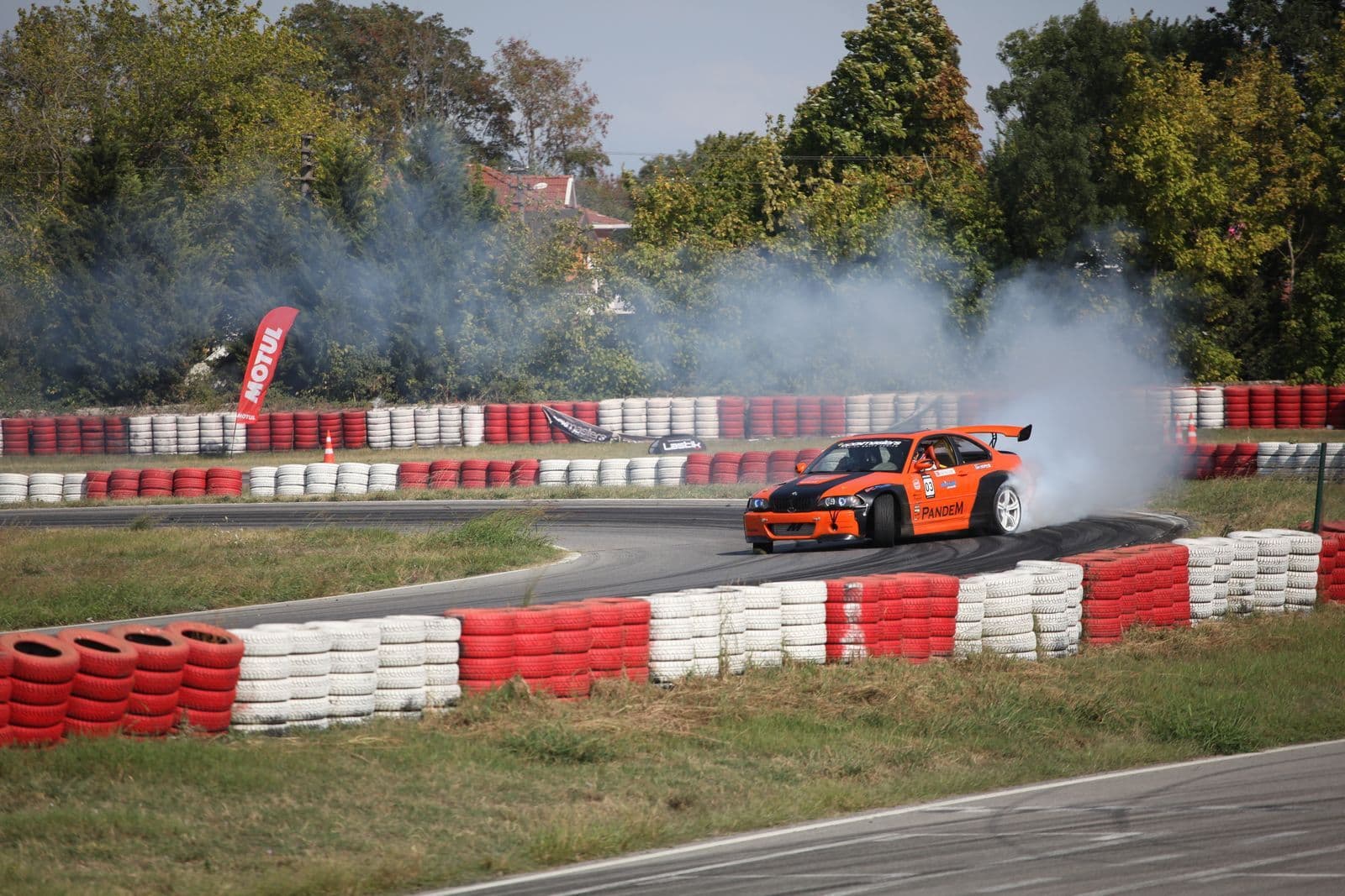 An orange race car with a rear wing drifts through a corner in a cloud of tyre smoke, between red and white tyre barriers.
