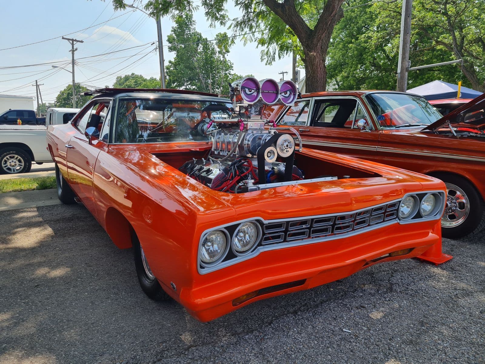 An orange classic muscle car with a chromed supercharger and twin carburettor scoops rising through its open bonnet, parked at an outdoor car show.