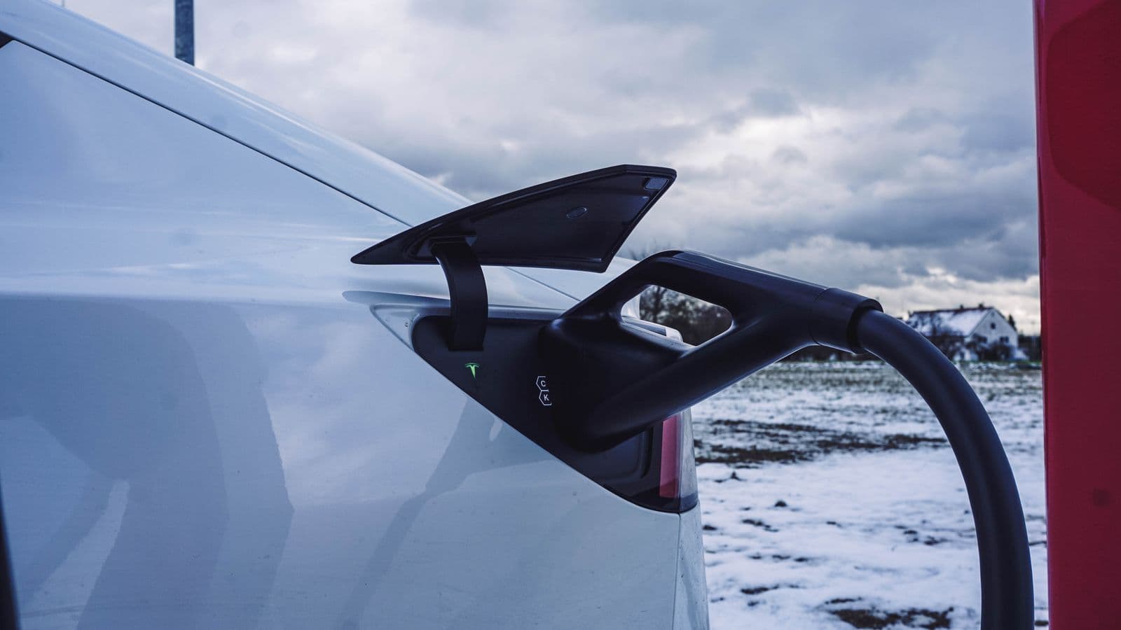 A charging cable plugged into the open charge port of a white electric car, with snowy fields and a grey sky behind.