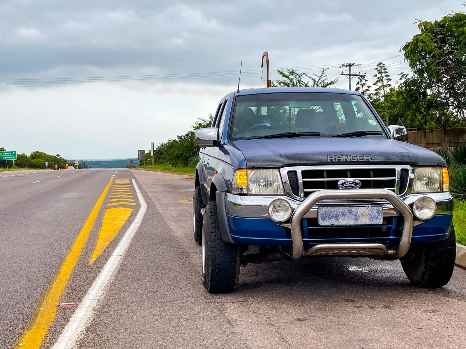 A blue dual-cab Ford Ranger ute with a chrome nudge bar parked on the shoulder of a two-lane highway under grey clouds.