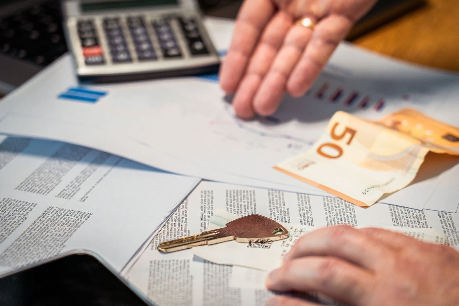 Hands over paperwork on a desk with a calculator, a banknote and a car key lying on the documents.