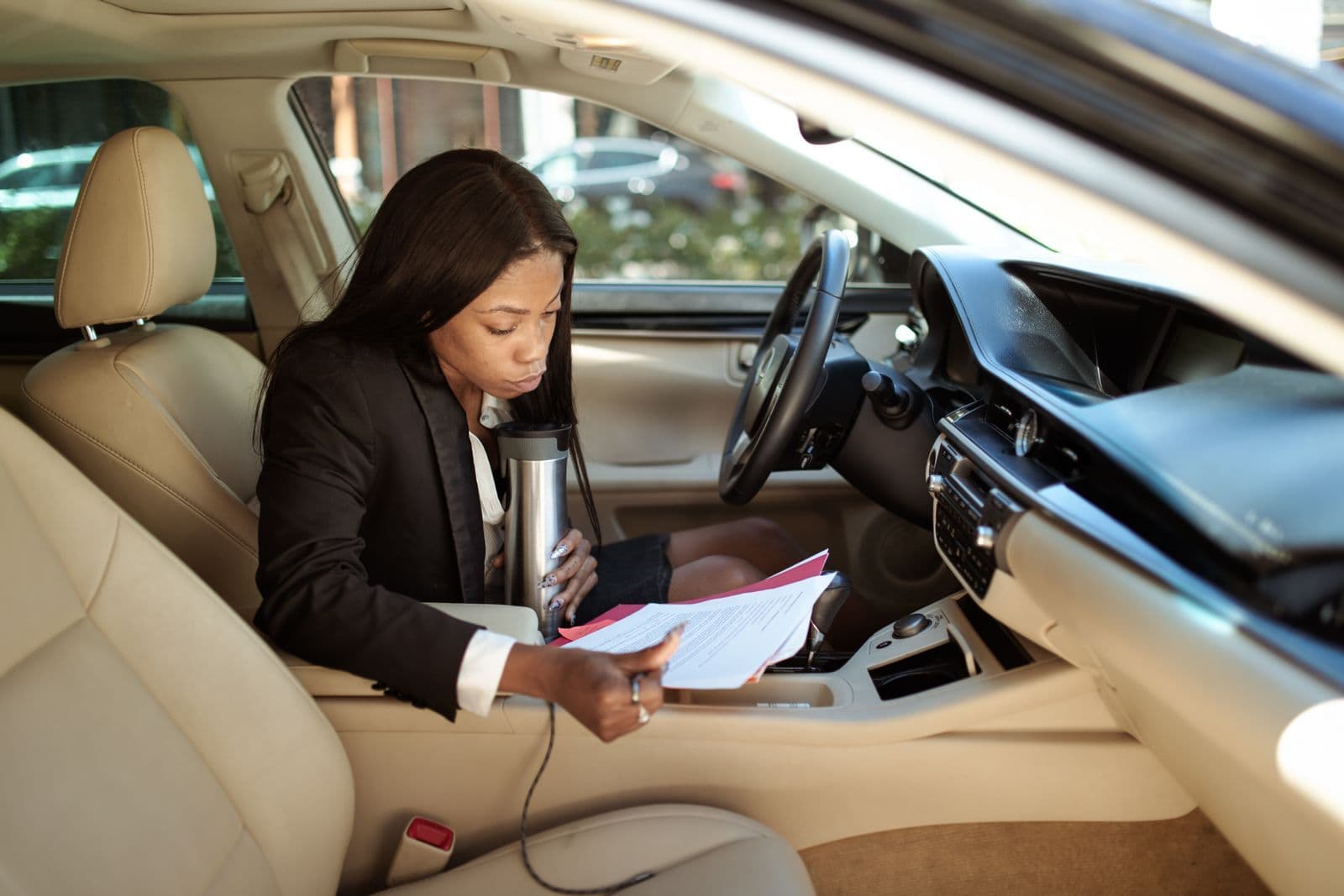A woman sitting in the driver's seat of a parked car, reading through a sheaf of printed paperwork resting on her lap.