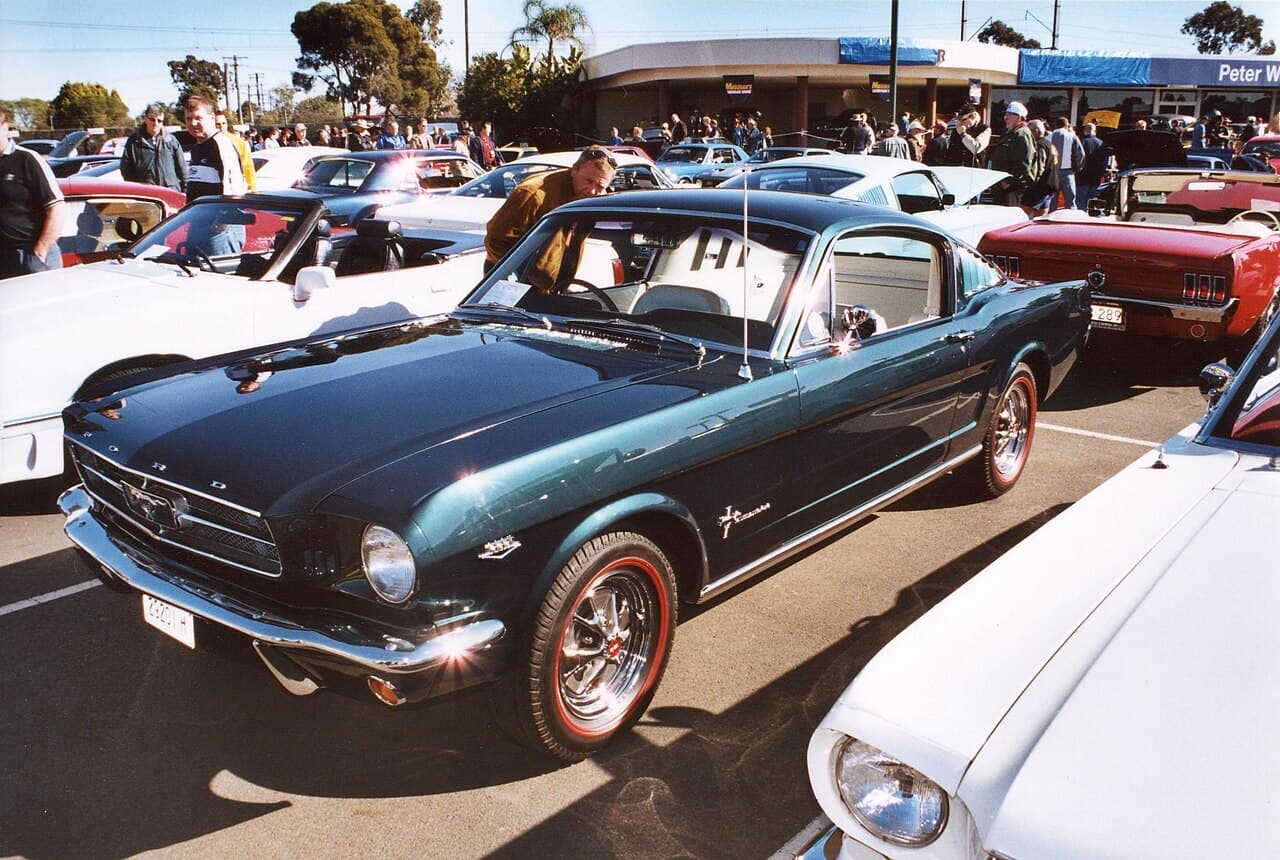 A dark green 1960s Ford Mustang fastback parked among other classic Mustangs at a crowded car show.
