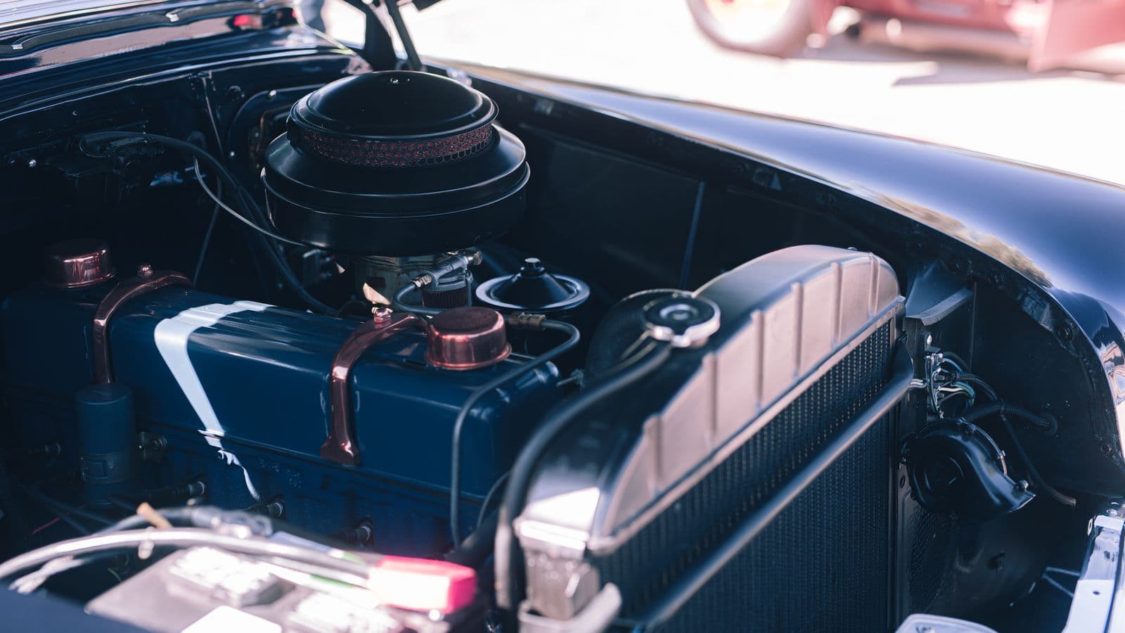 The engine bay of a vintage car with the bonnet raised, showing a blue-painted engine block, copper fittings and a round air cleaner beside the radiator.