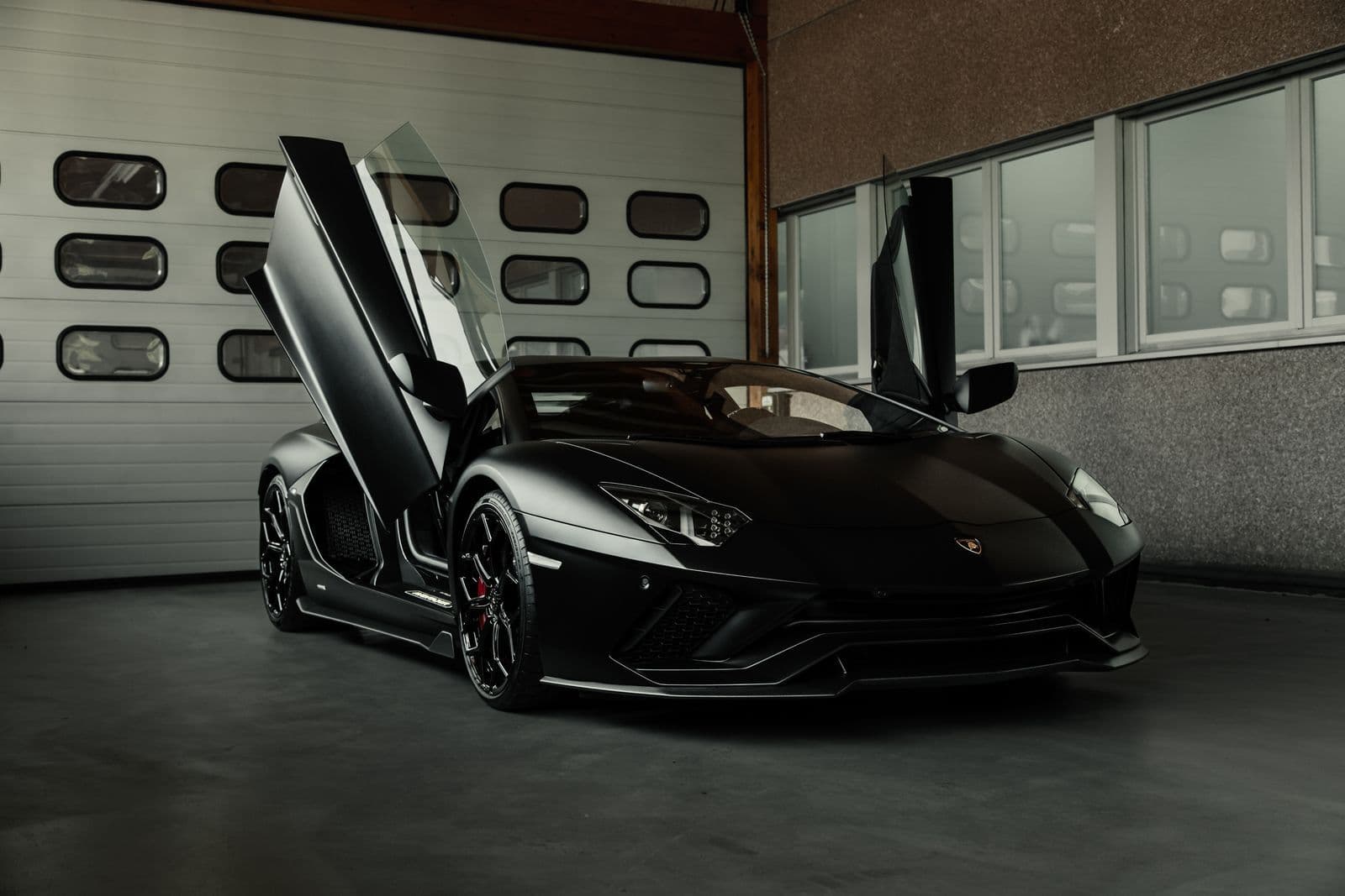 A matte black Lamborghini Aventador with its scissor doors raised, parked inside a garage in front of a roller door.