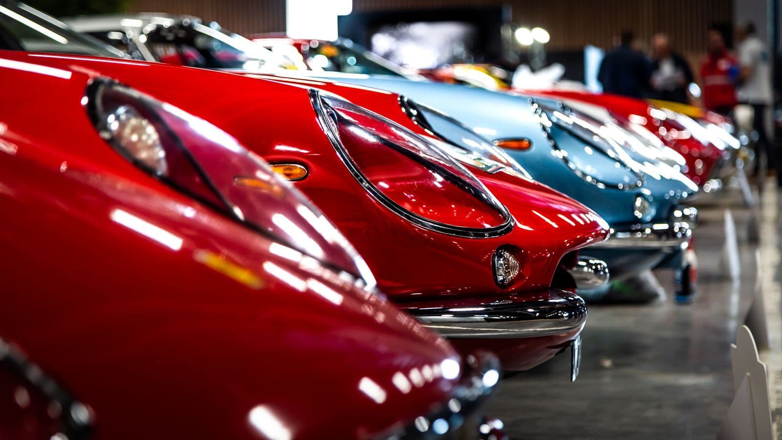 A row of polished classic sports cars lined up nose to tail on the floor of an indoor exhibition hall, with visitors browsing in the background.