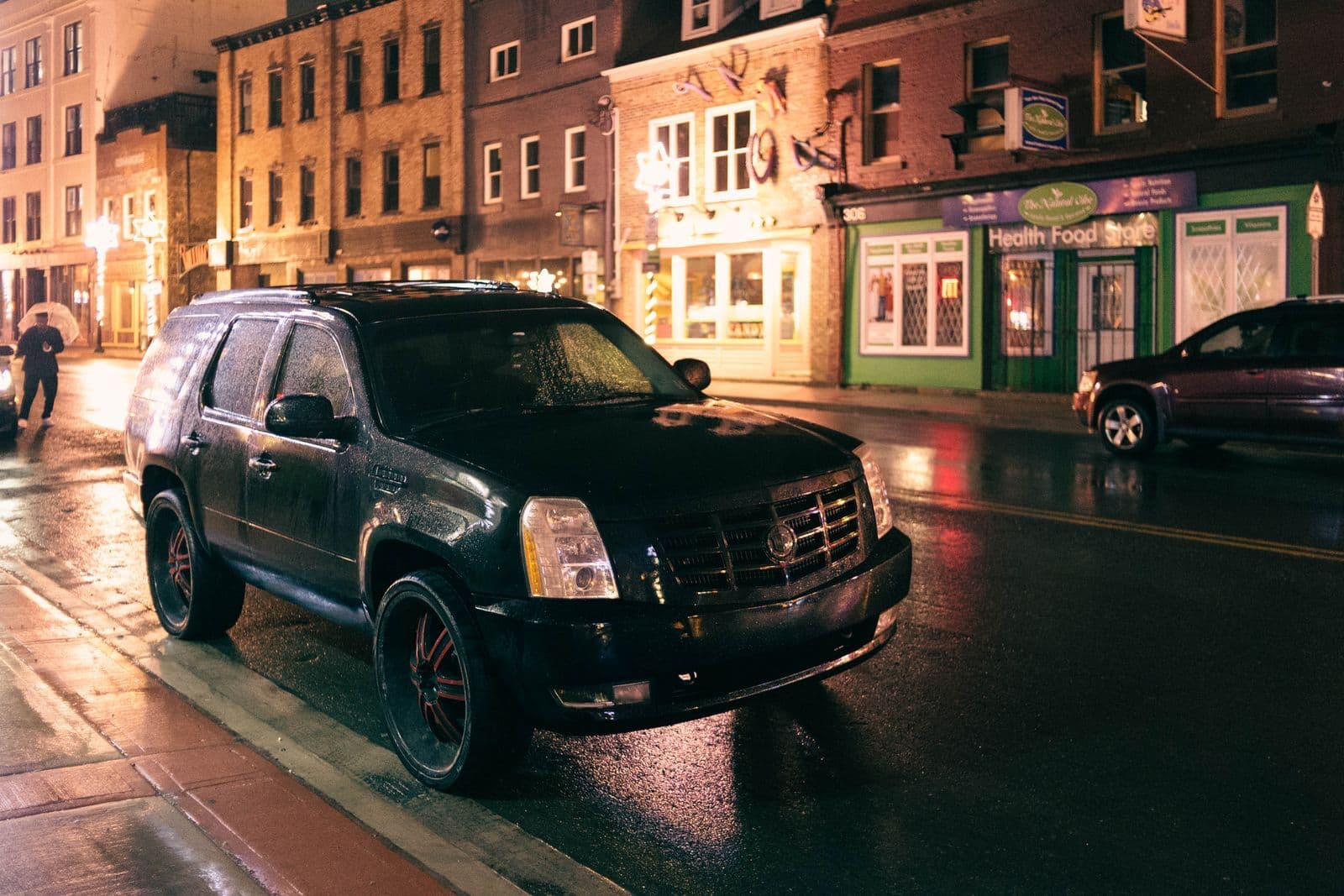 A black SUV parked alone on a wet street at night, lit by shopfront windows and streetlights on an empty road.