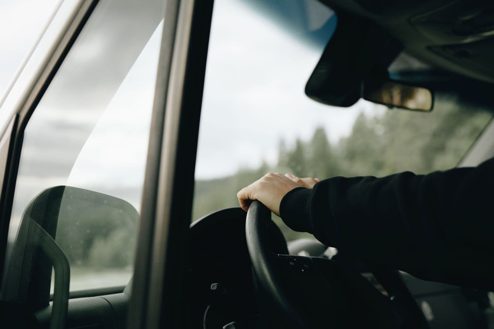 A driver's hand resting on the steering wheel of a car, seen from the passenger side, with a misty tree-lined road ahead through the windscreen.