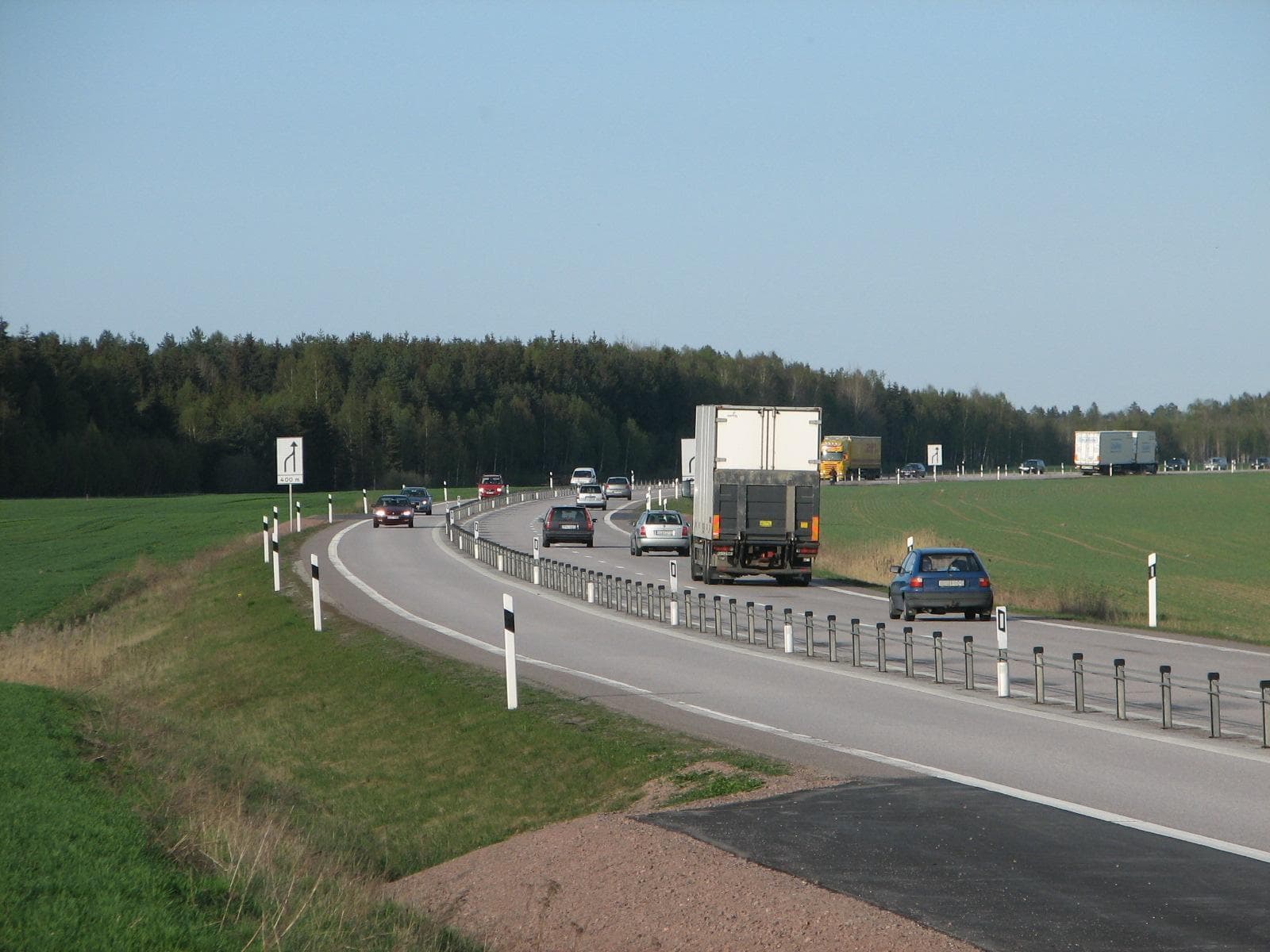 A Swedish rural road with two lanes in one direction and one in the other, separated by a steel cable barrier on posts, with cars and trucks passing green fields and forest.