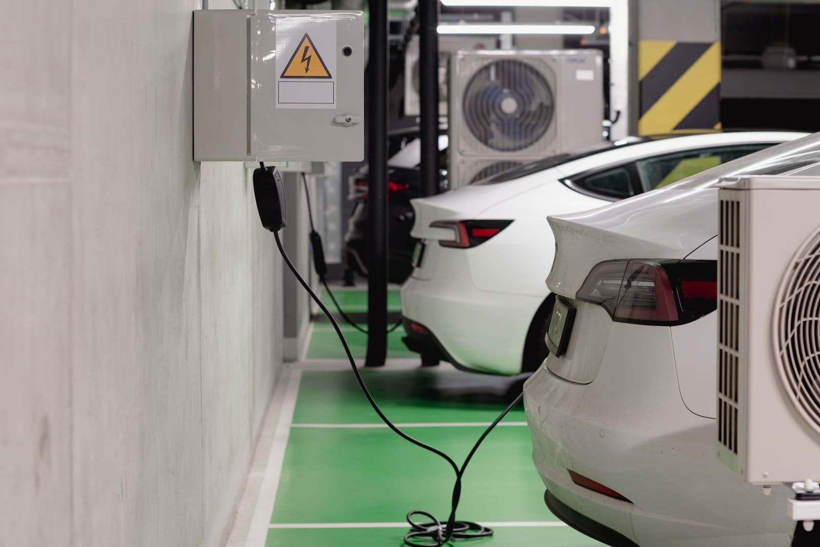 Two white electric cars plugged into a wall-mounted charger in a green-painted charging bay inside an underground car park.
