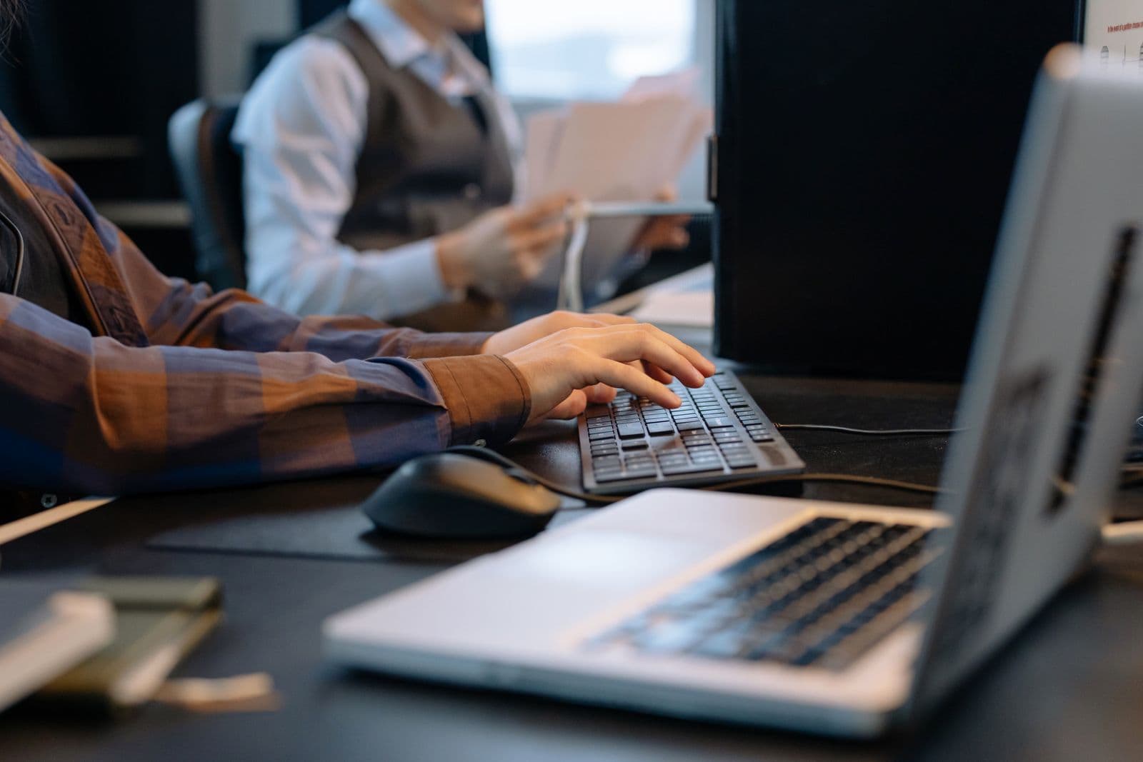 A person's hands typing on a keyboard at an office desk beside a laptop and mouse, with a colleague holding papers in the background.