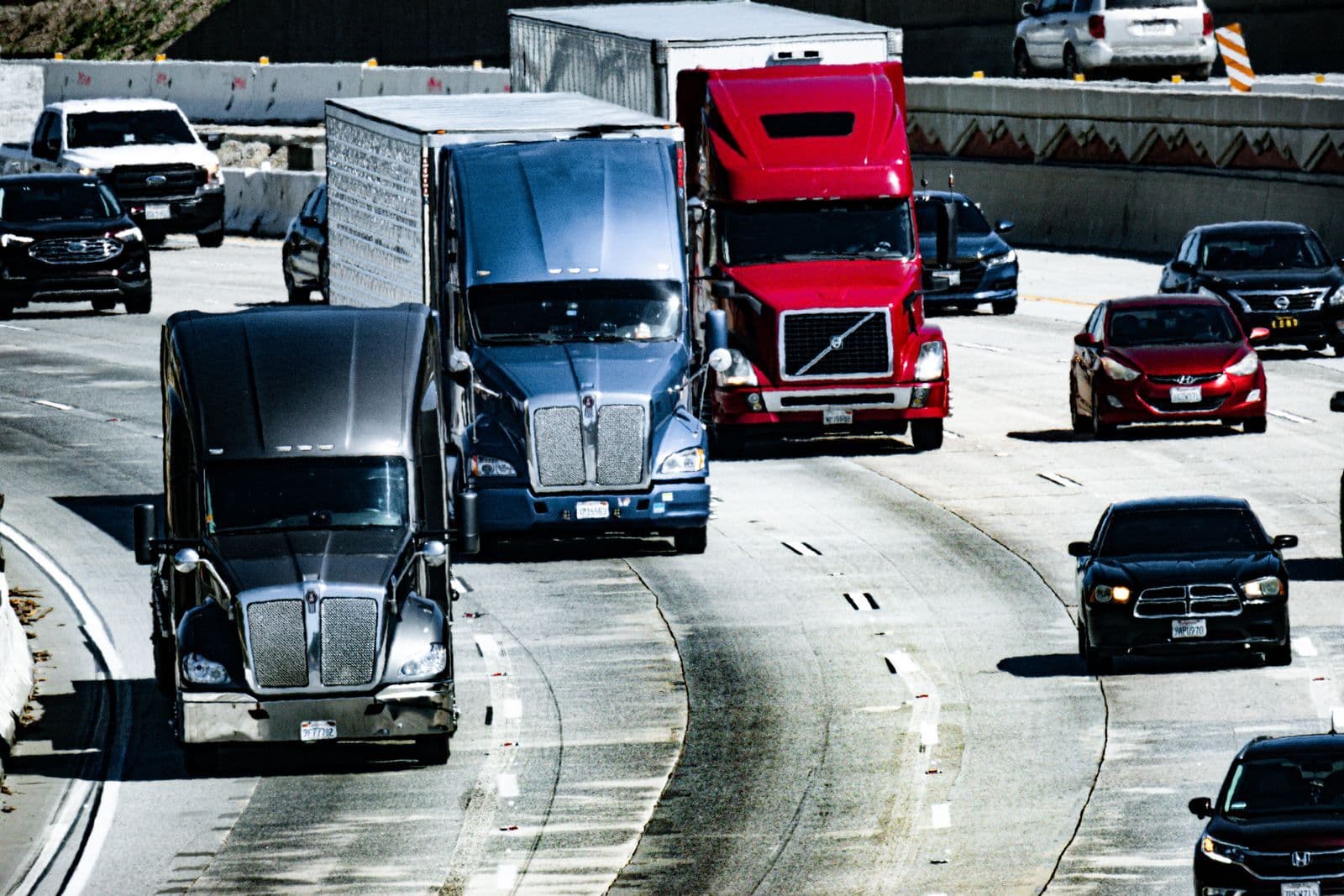 Semi-trailers and cars sharing several lanes of a busy multi-lane highway in daylight.