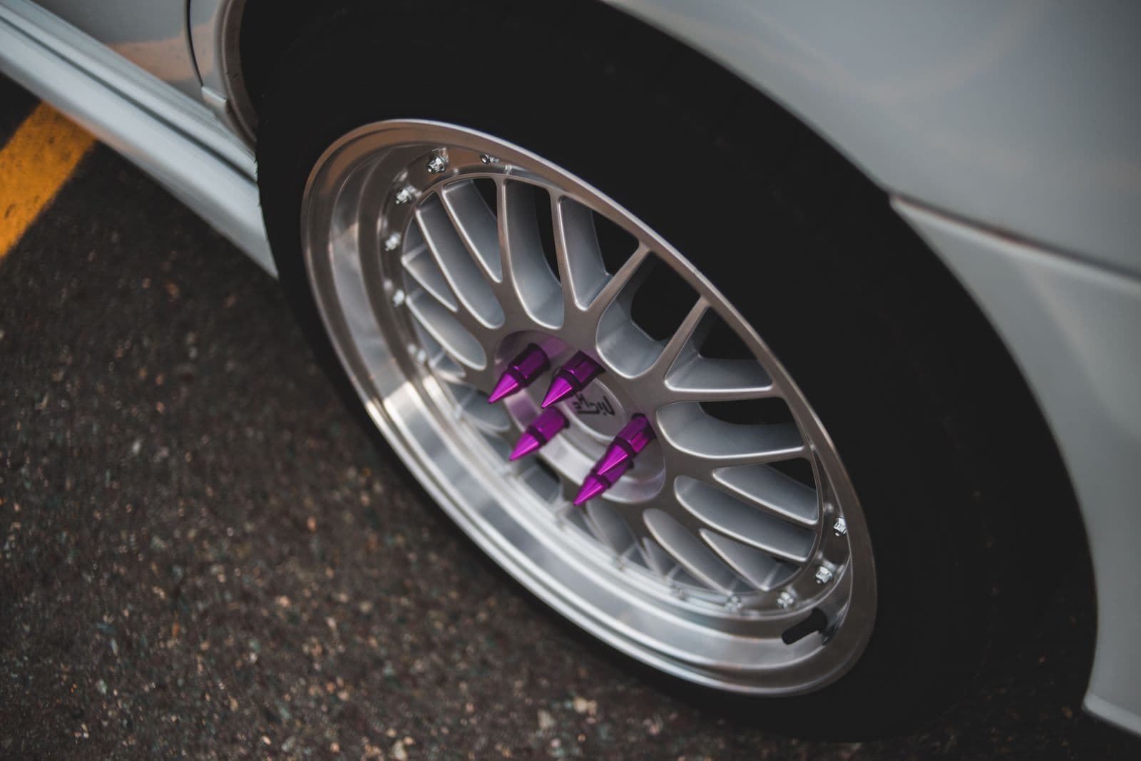 A polished aftermarket mesh-style alloy wheel on a silver car, fastened with purple spiked wheel nuts.