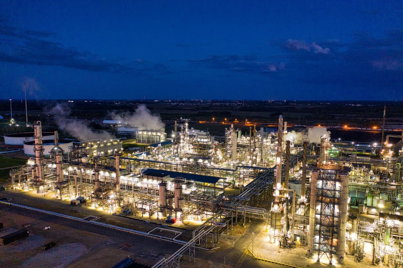 An aerial view of an oil refinery lit up at night, with steam rising from its towers.