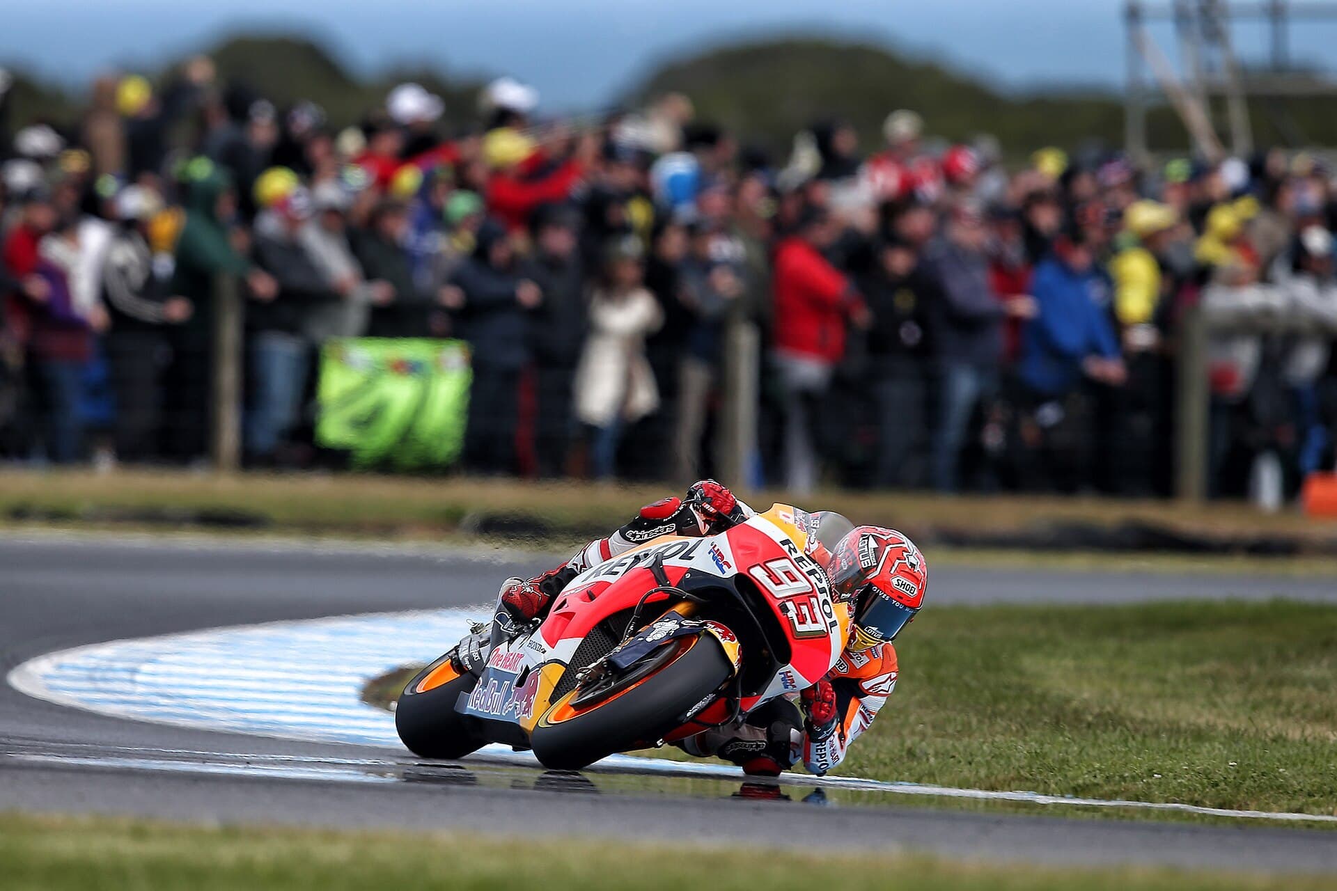 A Repsol Honda MotoGP rider leans hard through a corner at Phillip Island with a packed crowd on the grass bank behind.