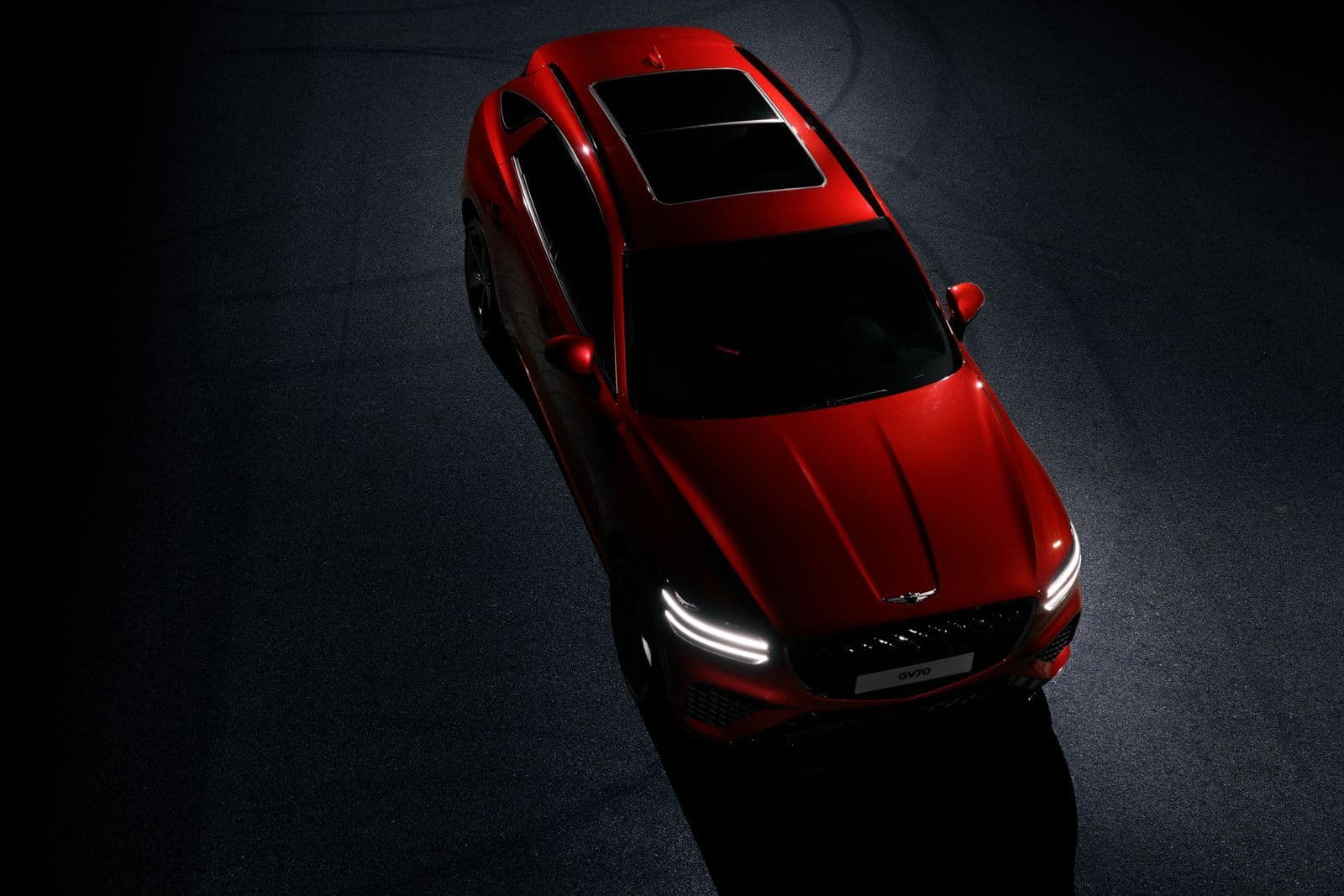A red SUV seen from above under a single spotlight on a dark showroom floor, its bonnet and glass roof catching the light.