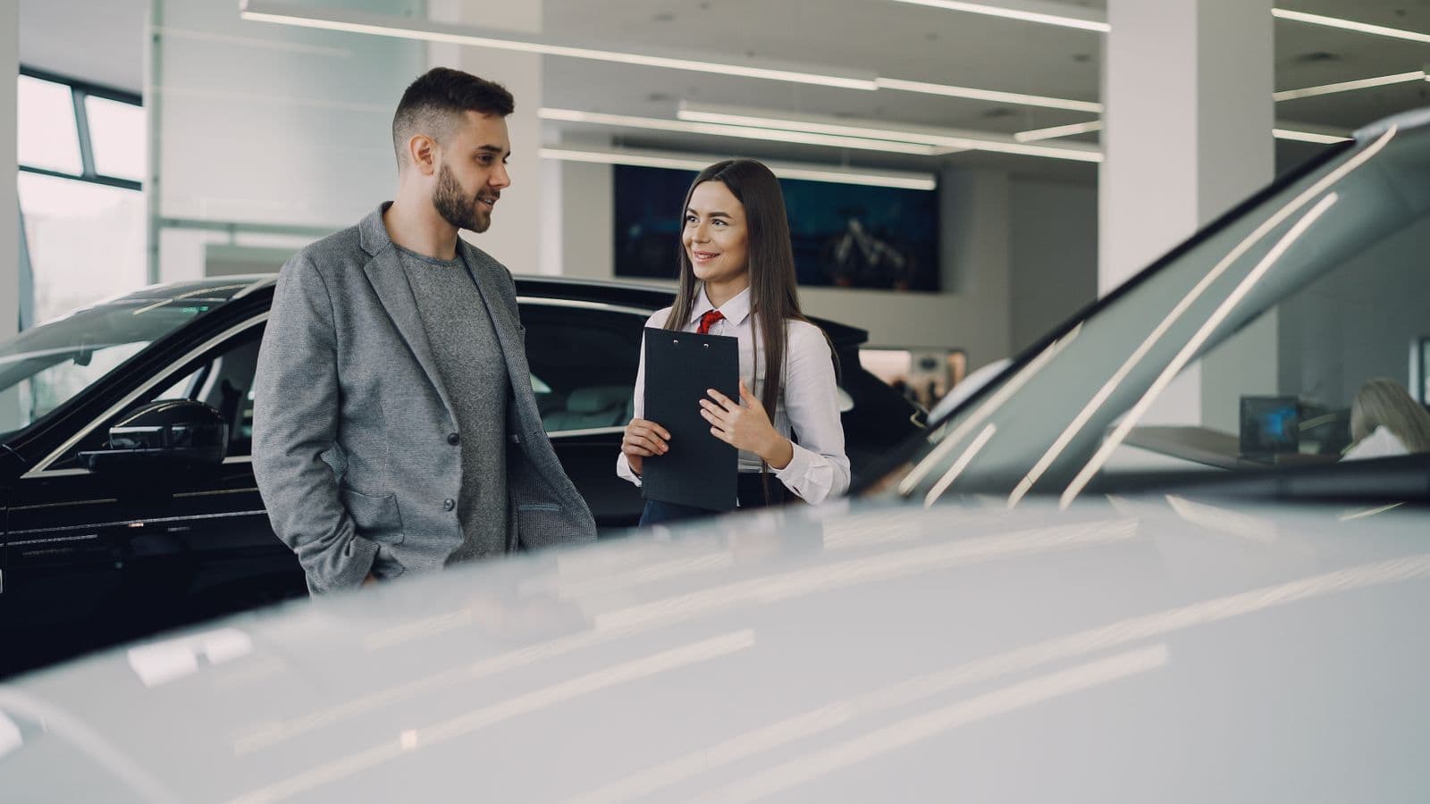 A saleswoman holding a clipboard talks with a customer between two cars on a bright dealership floor.