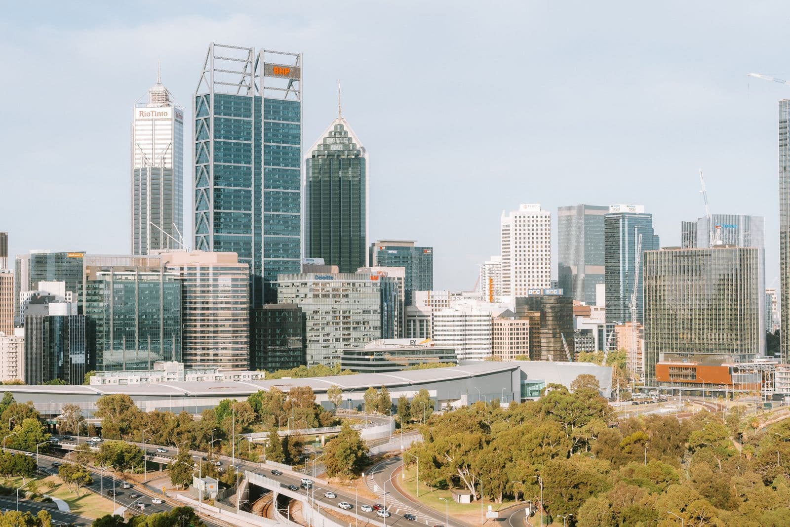 The Perth city skyline above a freeway interchange carrying traffic past parkland.