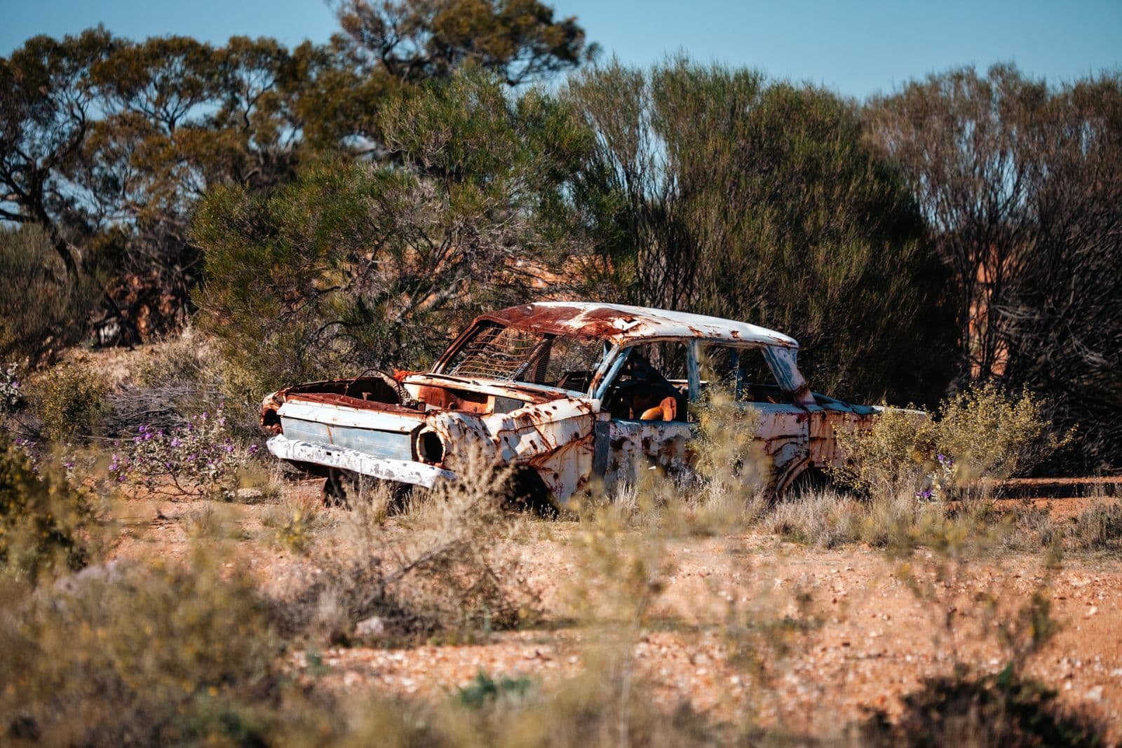 A stripped, rusting car abandoned among low scrub and red dirt in the Australian bush.