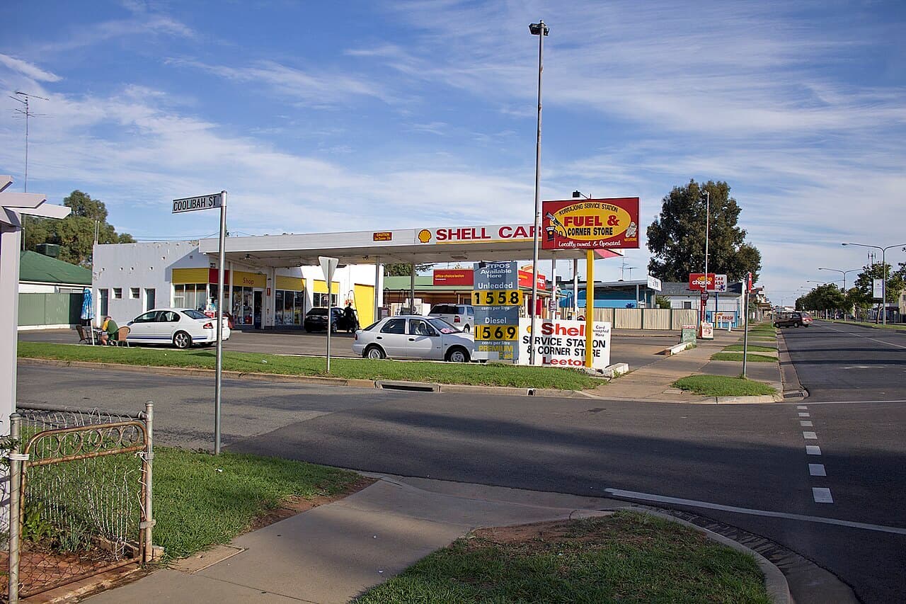 A small Shell service station on a quiet street corner in a country town, with a price board, a forecourt and two parked cars.