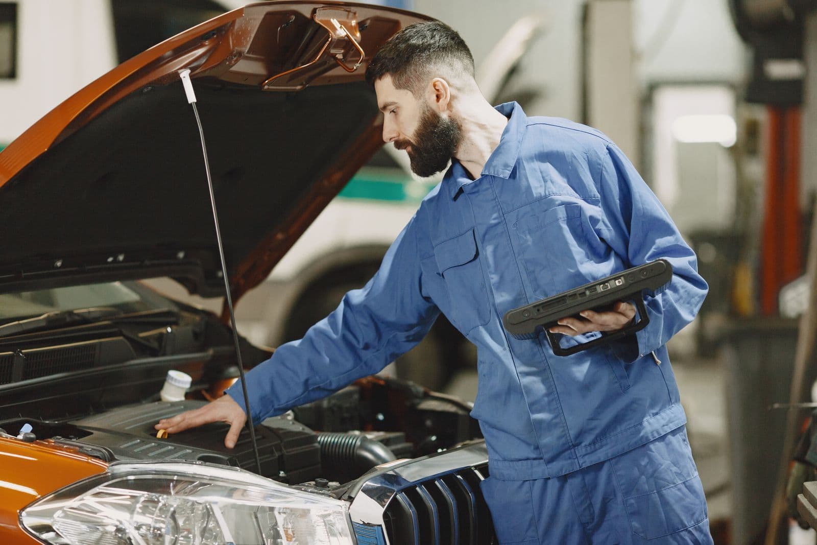 A mechanic in blue overalls holding a tablet leans over the open engine bay of an orange car in a workshop.
