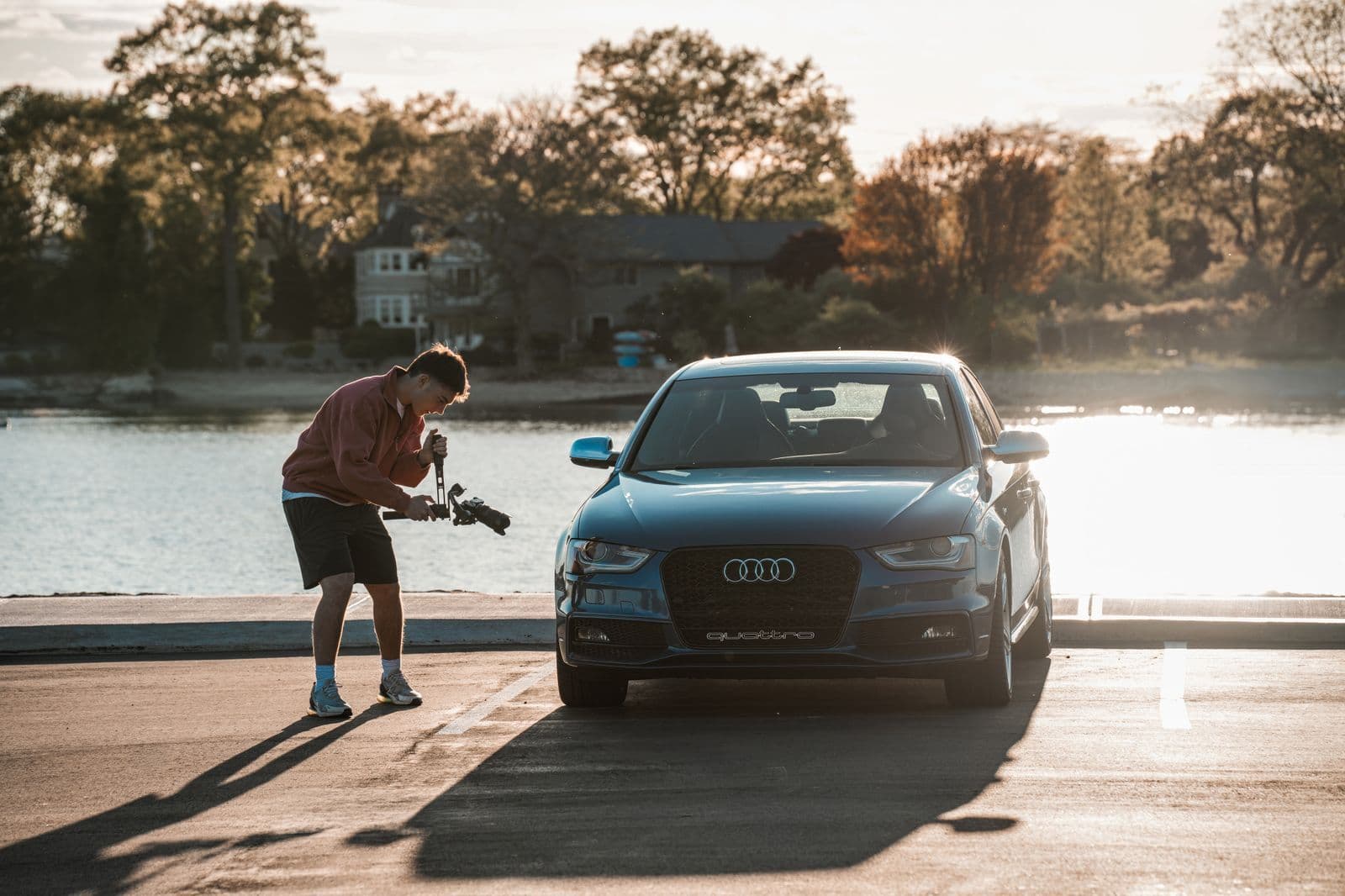 A person crouches with a camera to photograph the front of a dark sedan parked by the water at sunset.