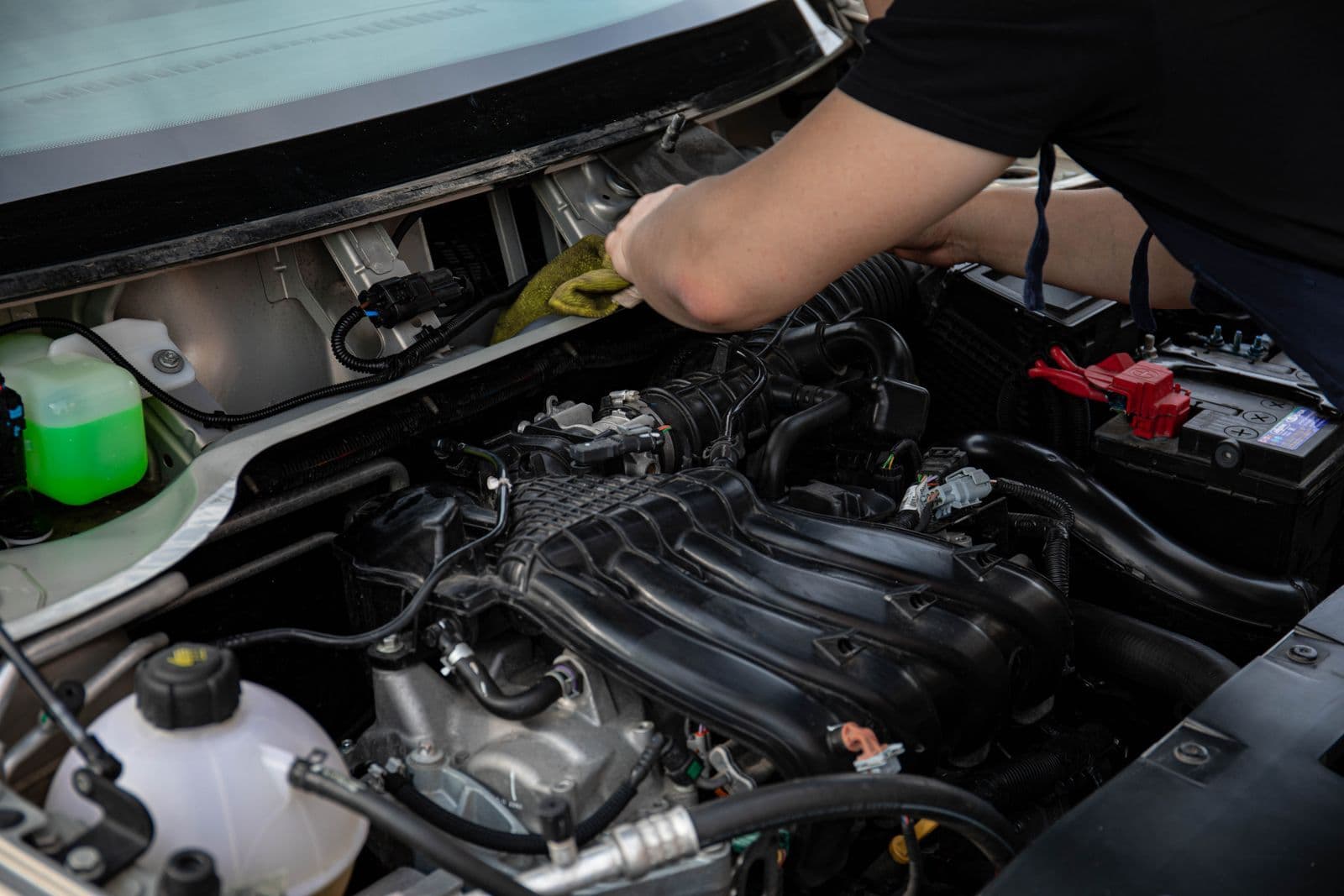 A person's arm reaching into an open car engine bay with a cloth, beside the coolant reservoir and battery.