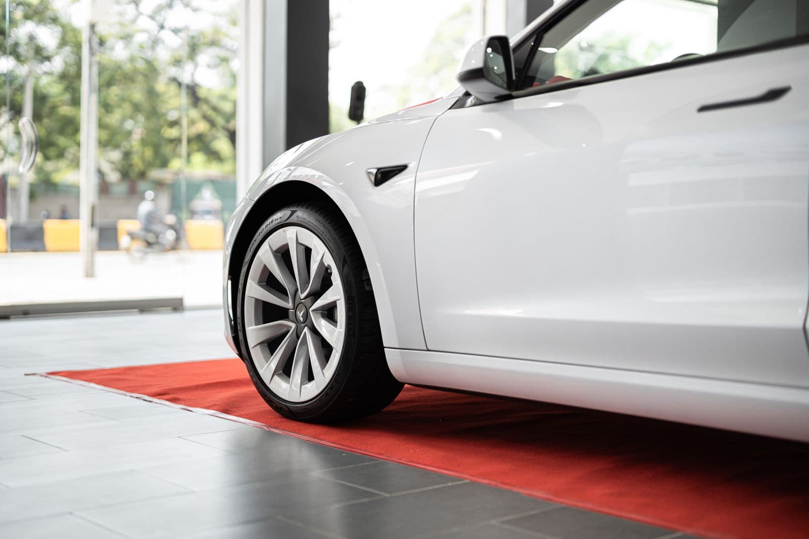 A white electric sedan standing on a red carpet just inside a glass showroom door, photographed low beside its front wheel.