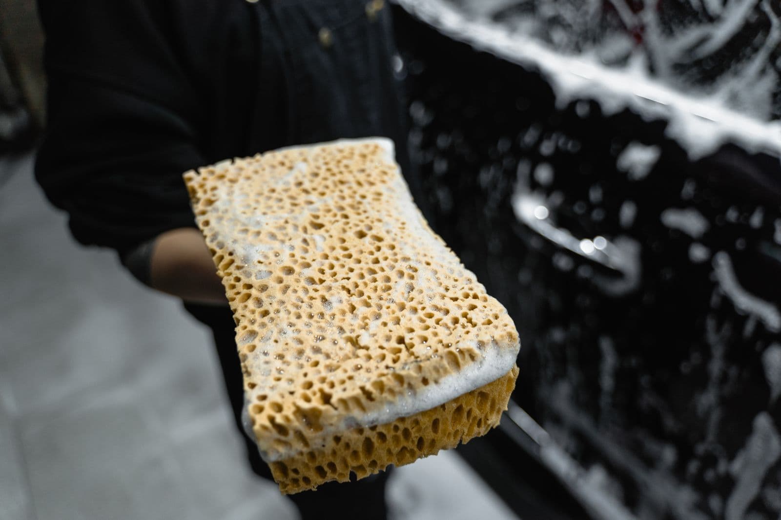 A hand holding a soapy sponge beside a black car covered in suds.