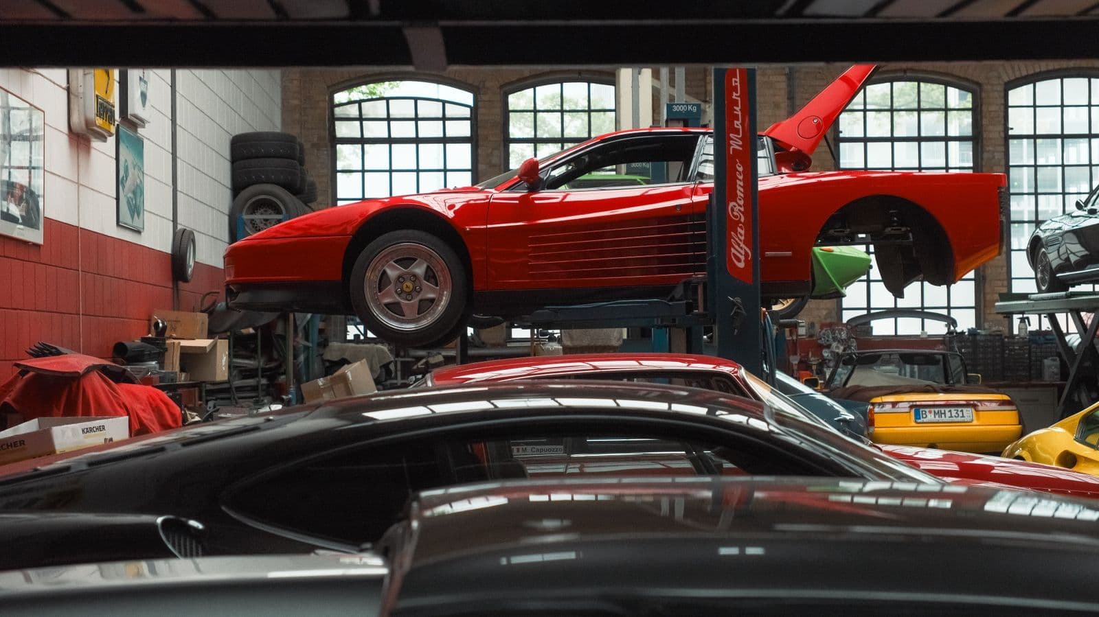 A red mid-engined sports car raised on a two-post hoist in a specialist workshop, with other low-slung cars parked below it.