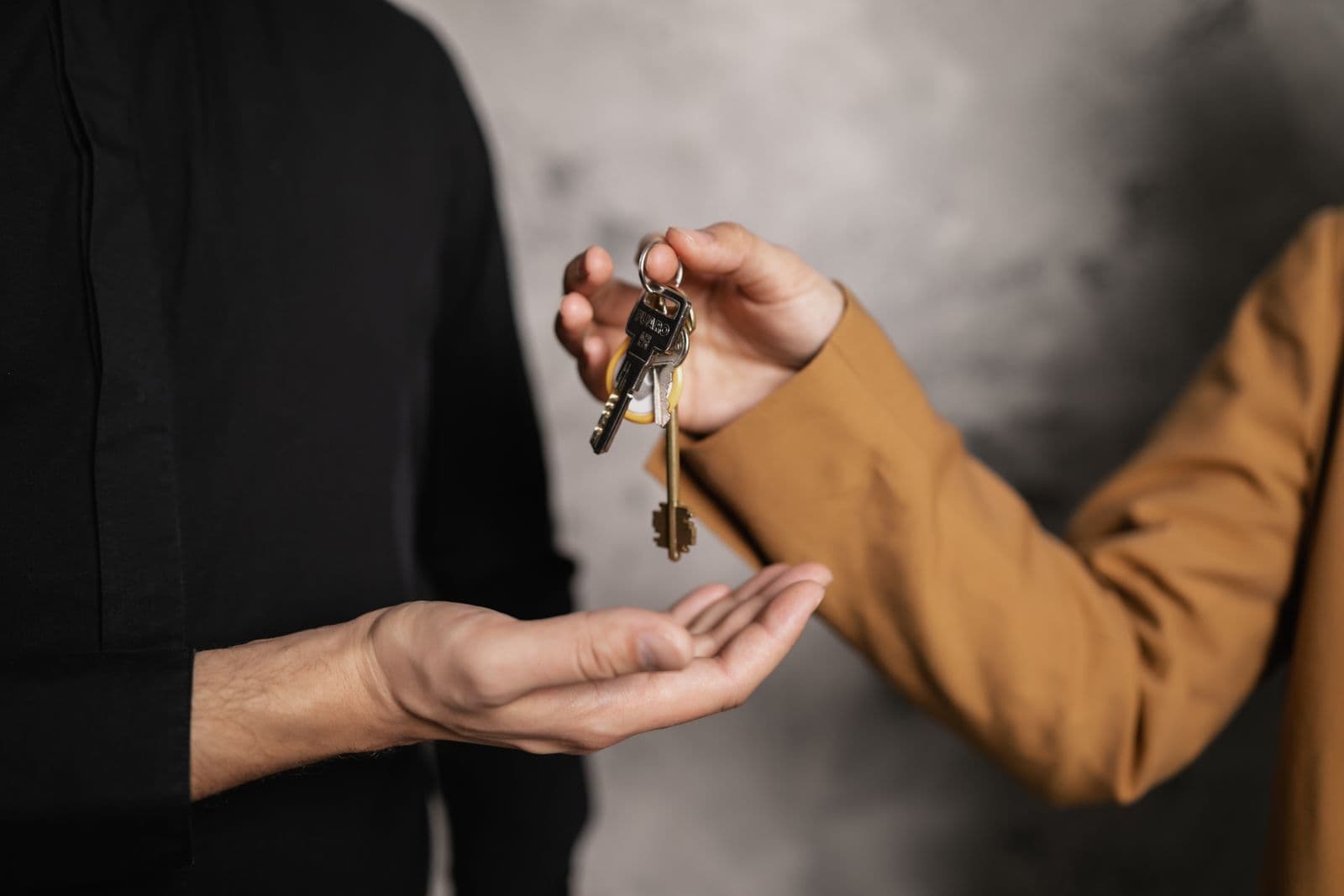 One person drops a bunch of keys into another person's open palm against a plain grey wall.