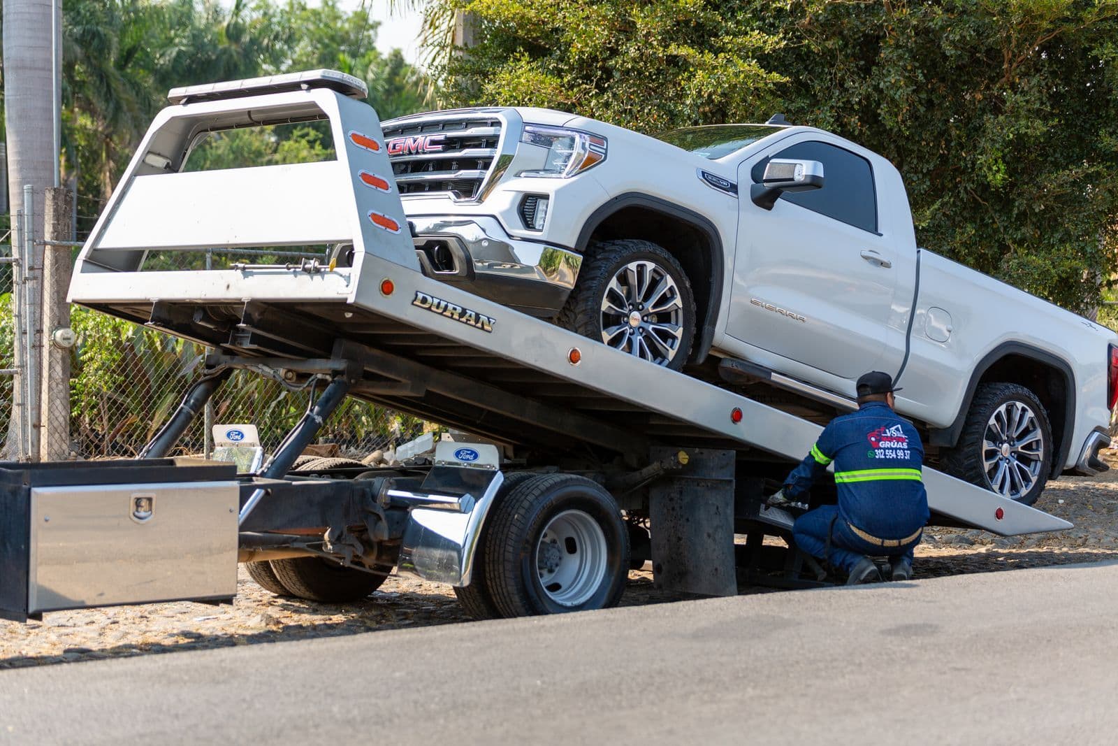 A white dual-cab pickup winched up onto the tilted deck of a flatbed tow truck while an operator crouches at the rear.