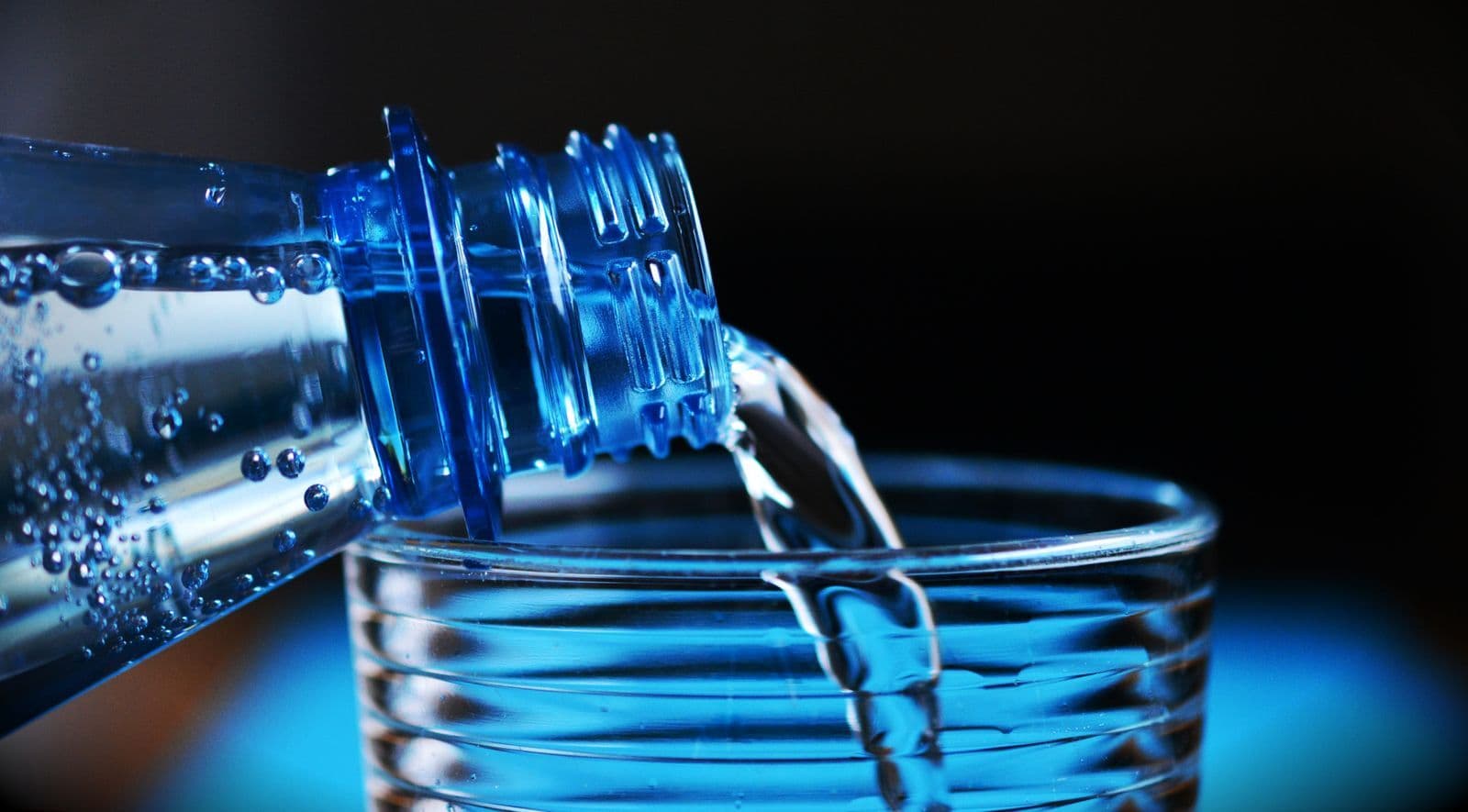 Water pouring from a plastic bottle into a ribbed glass, lit blue against a dark background.