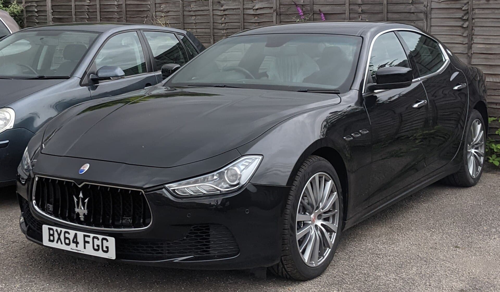 A black Maserati Ghibli sedan parked on a gravel lot beside another car, seen from the front three-quarter angle.