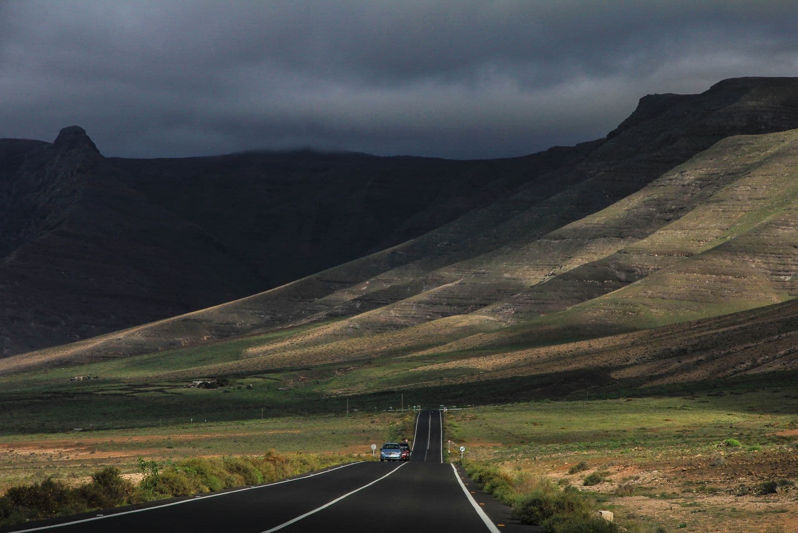 A lone car drives along an empty country road toward steep, grassy hills under dark storm clouds.