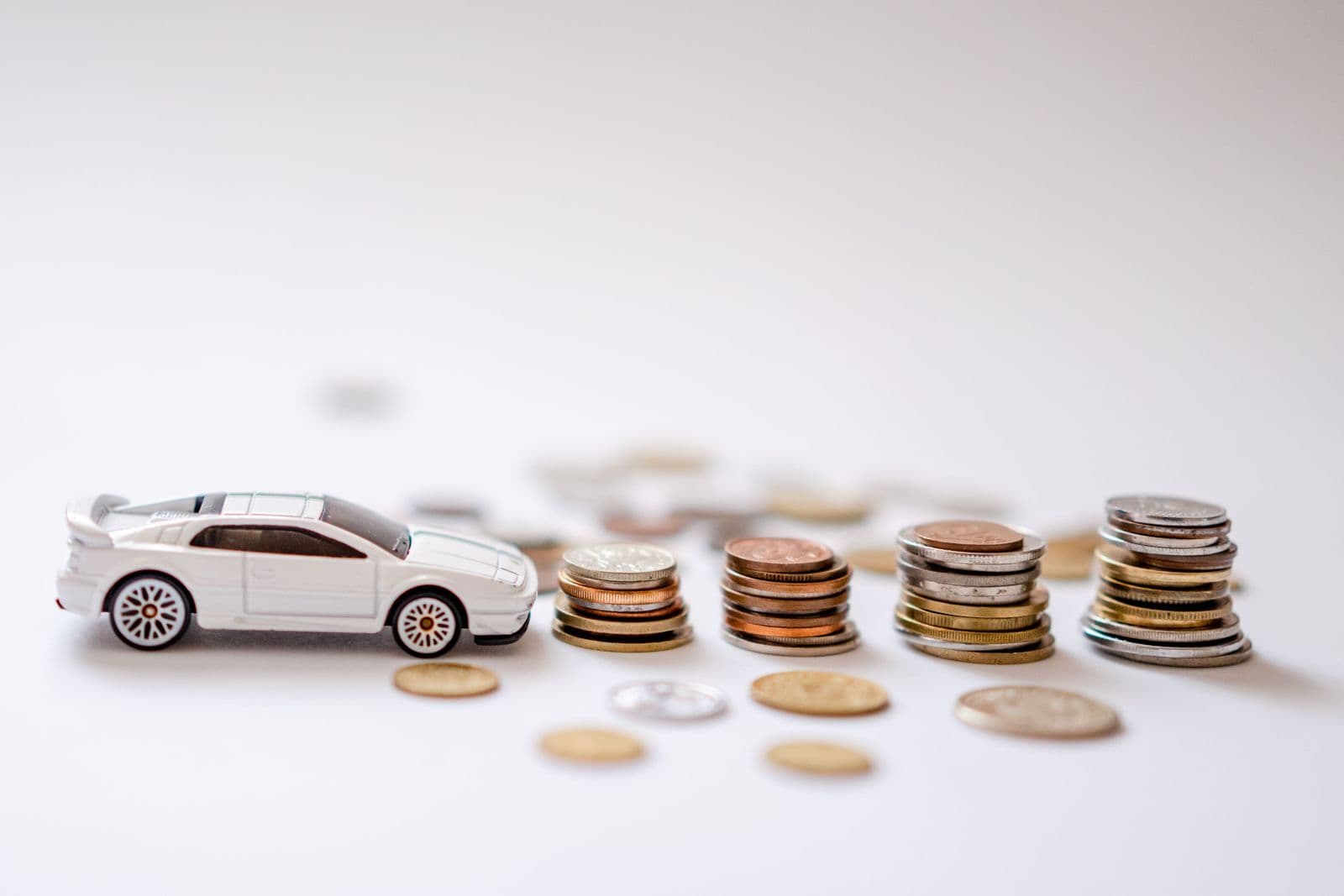 A small white model car on a white surface beside several stacks of coins.