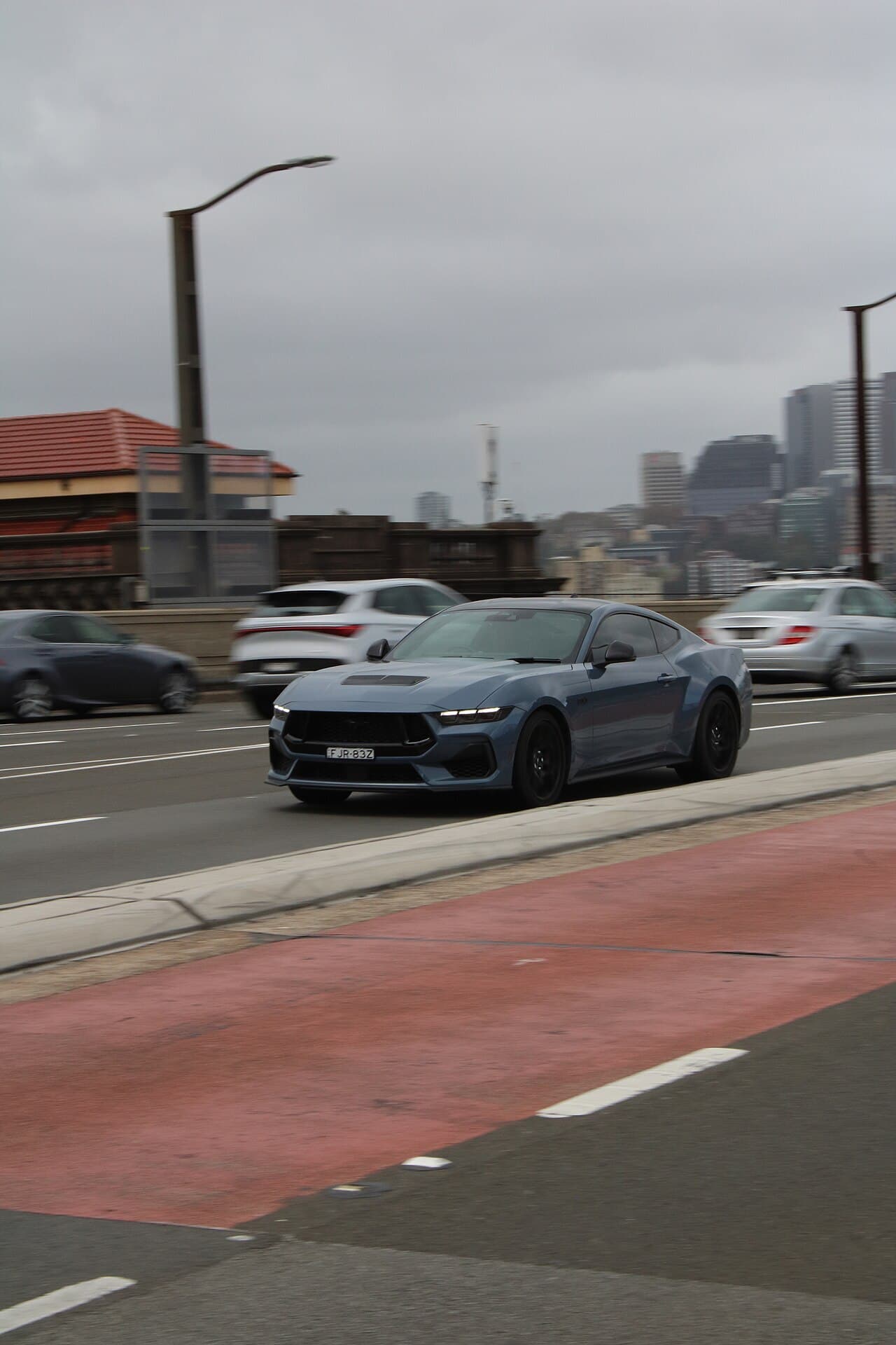 A blue-grey current-generation Ford Mustang coupe driving across the Sydney Harbour Bridge with the city skyline behind.