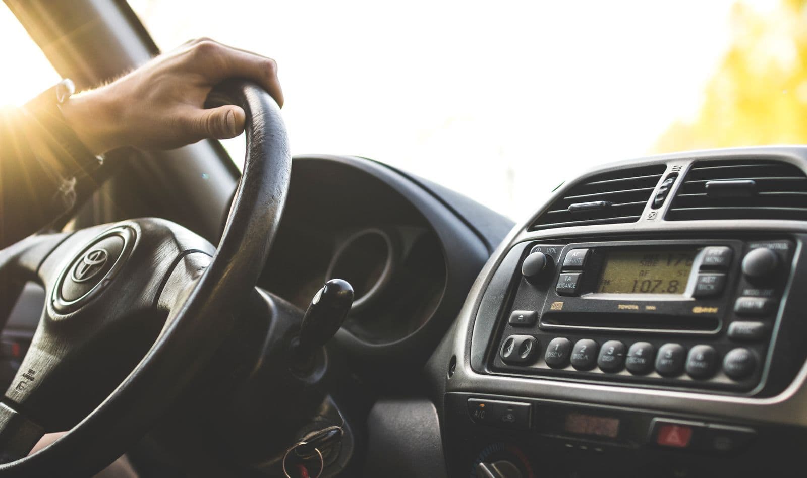 A driver's hand rests on the steering wheel of a car, with the dashboard and radio lit by bright sunlight through the windscreen.