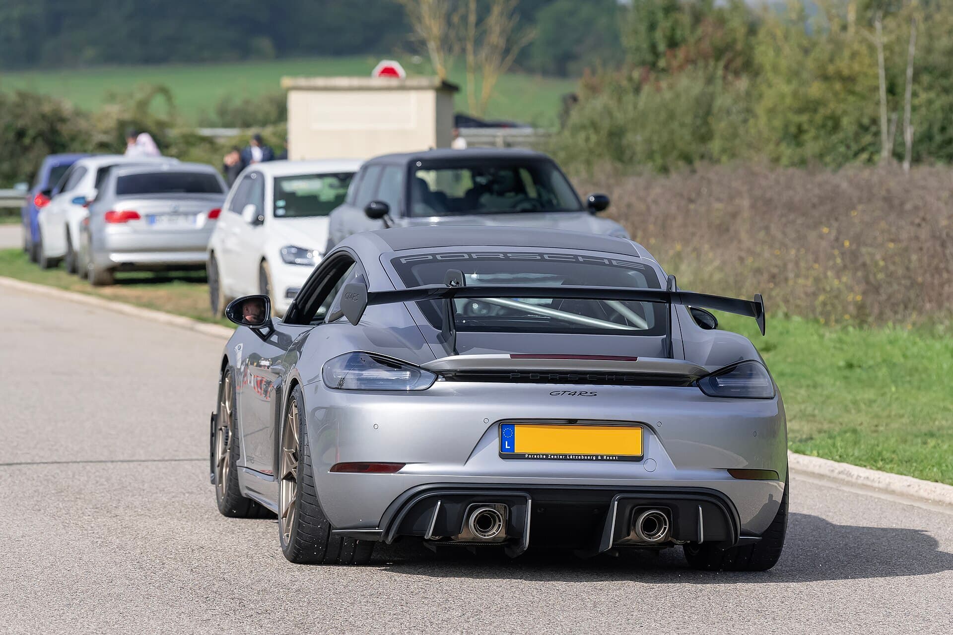 A silver Porsche 718 Cayman GT4 RS with a large rear wing, seen from behind on a road with parked cars beyond.
