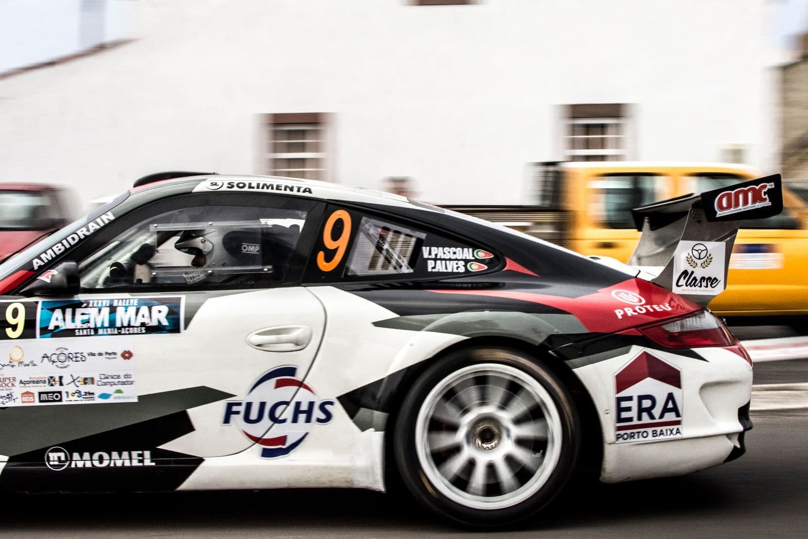 A white, black and red Porsche 911 race car with sponsor decals and a rear wing, blurred by speed as it passes a building.