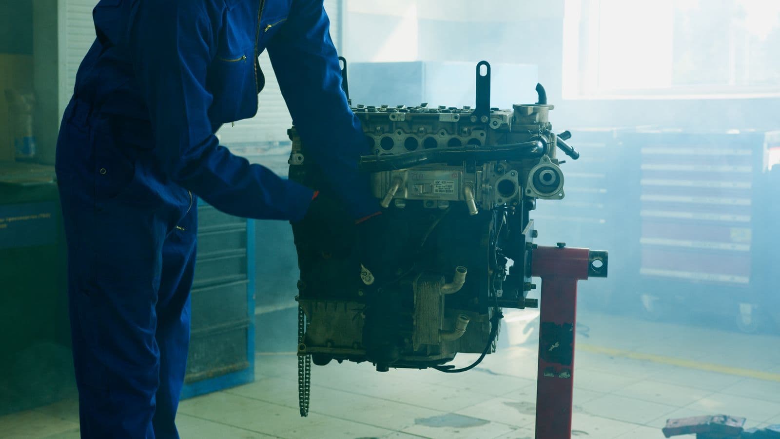 A mechanic in blue overalls steadies a bare engine block mounted on a red engine stand in a hazy workshop.